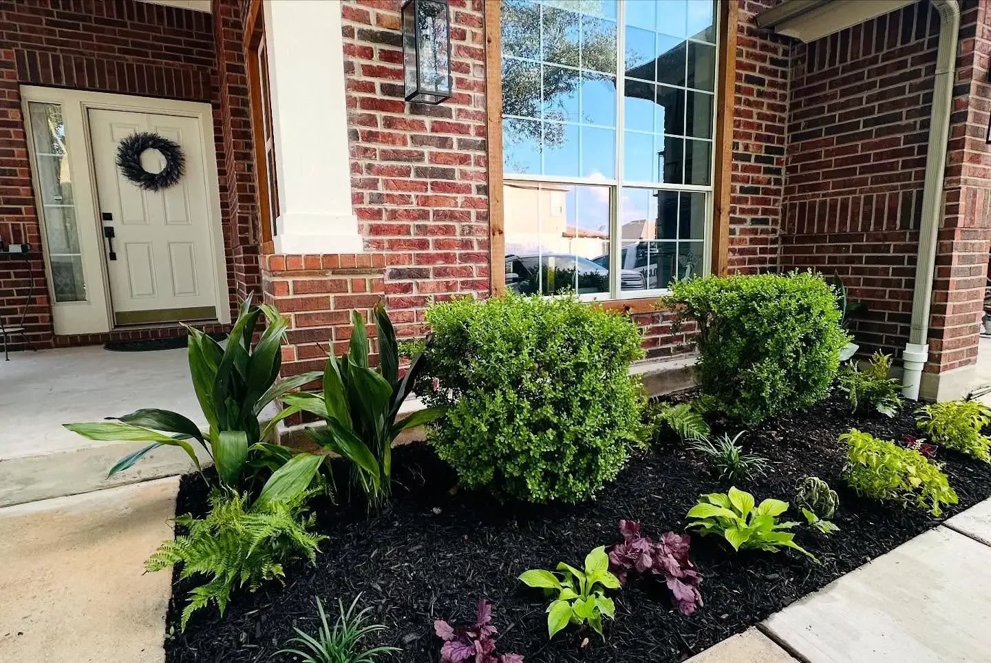 Close-up view of a well-maintained garden bed with green shrubs, leafy plants, and dark mulch in front of a brick house with a large window and a white door with a wreath.