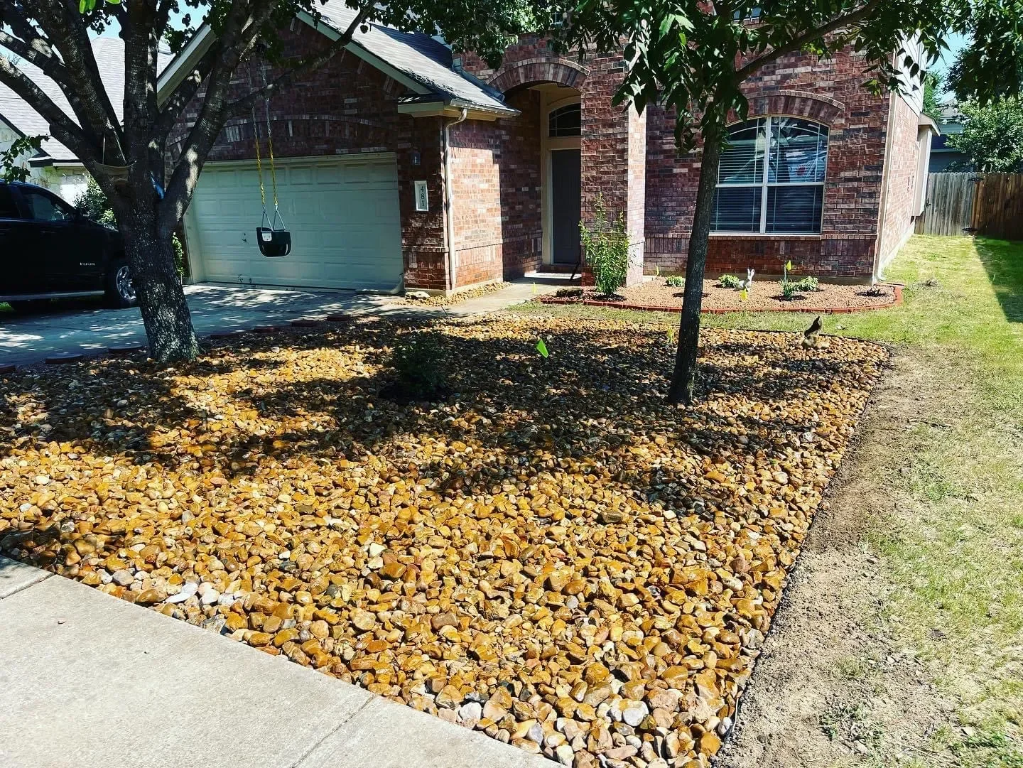 Front yard of a house with two trees, a rocky landscape, and a brick house with large windows and a garage.