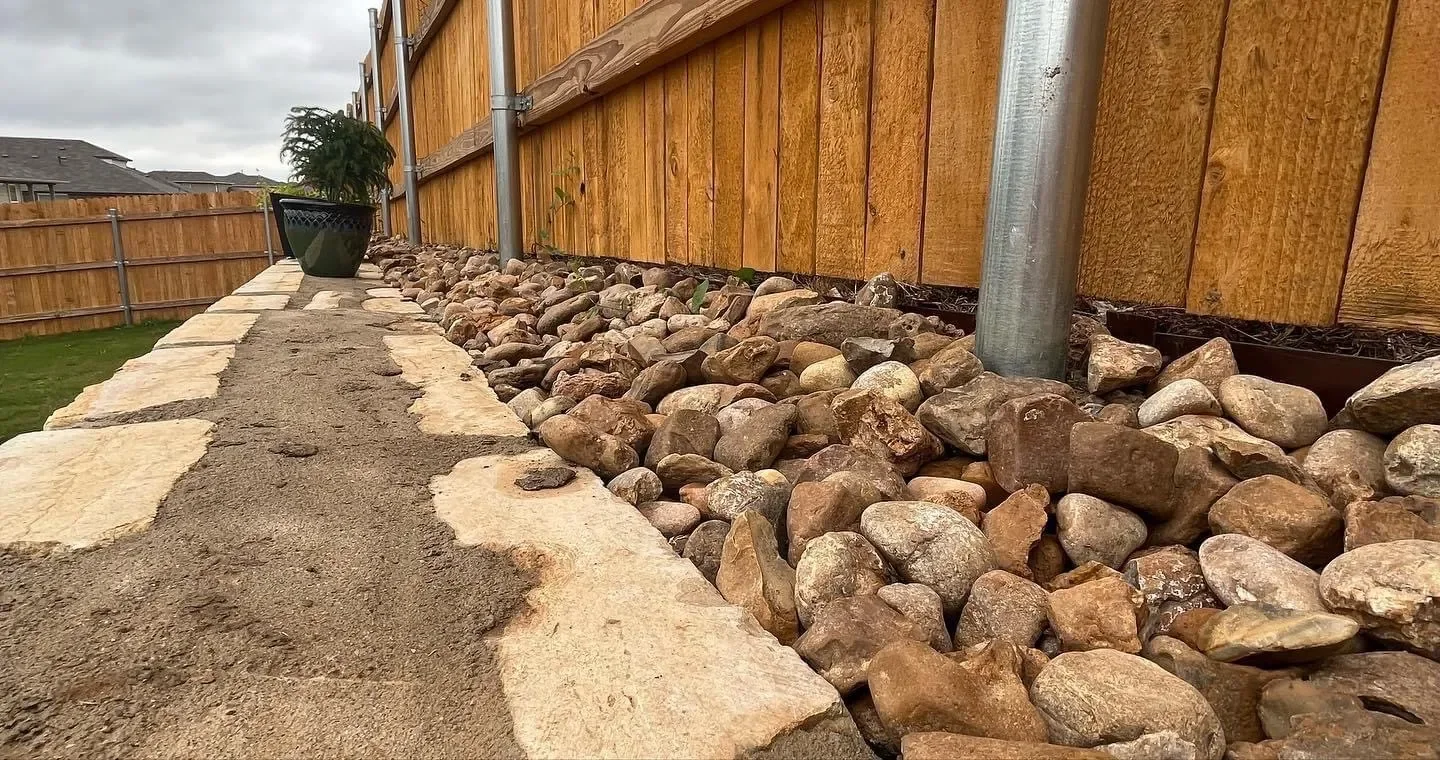 A pathway made of flat stones and dirt alongside a wooden fence with rocks and a potted plant beside the fence.