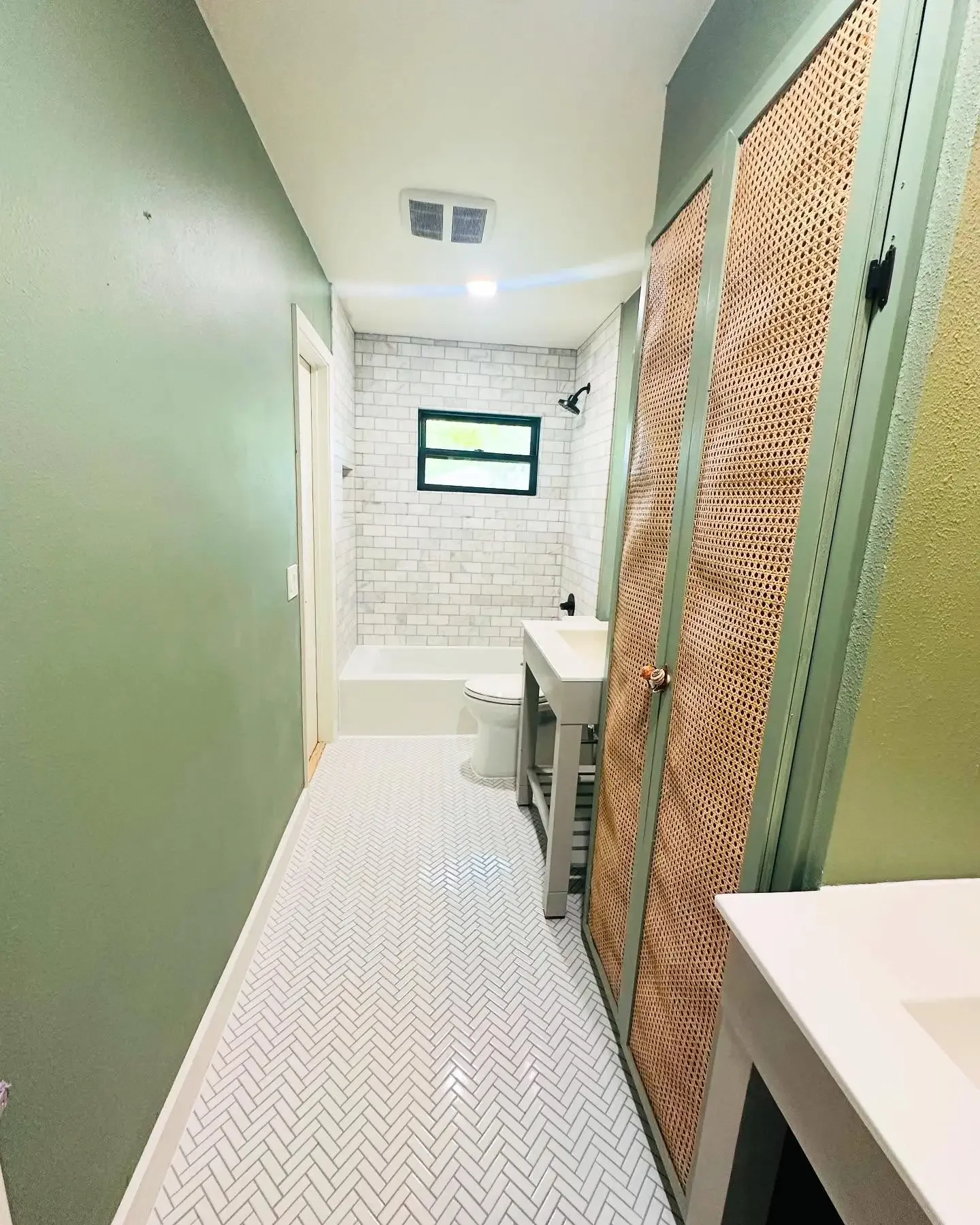 Bathroom with green walls, white herringbone tile floor, walk-in shower with white subway tile, small black-framed window, and a vanity with a vessel sink. There is a wooden cabinet with rattan-style doors on the right side.