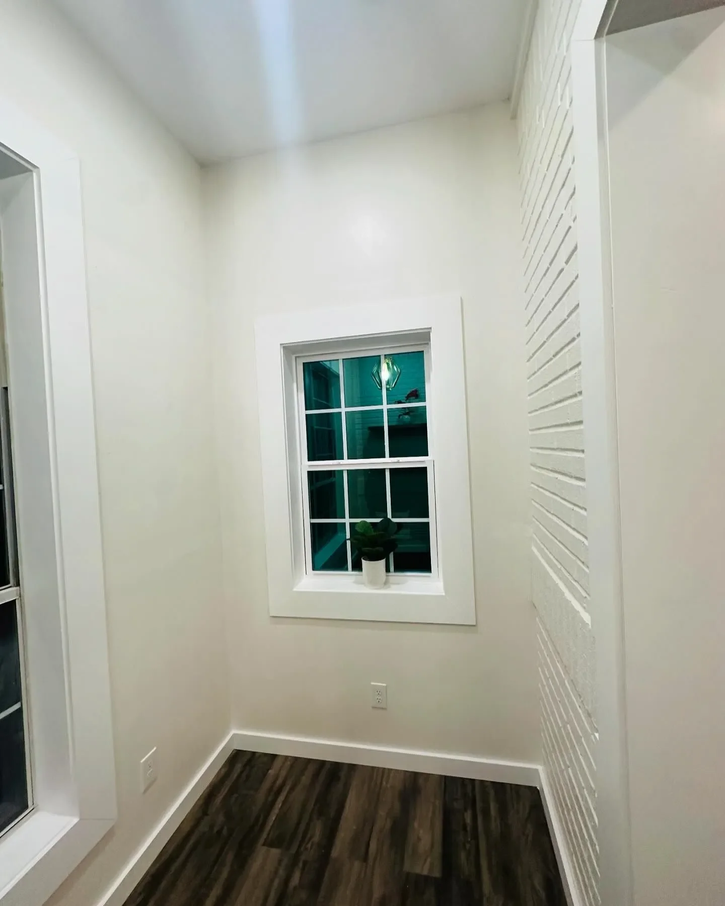 A small white room with a window holding a potted plant, hardwood flooring, and white walls with a textured section on the right.