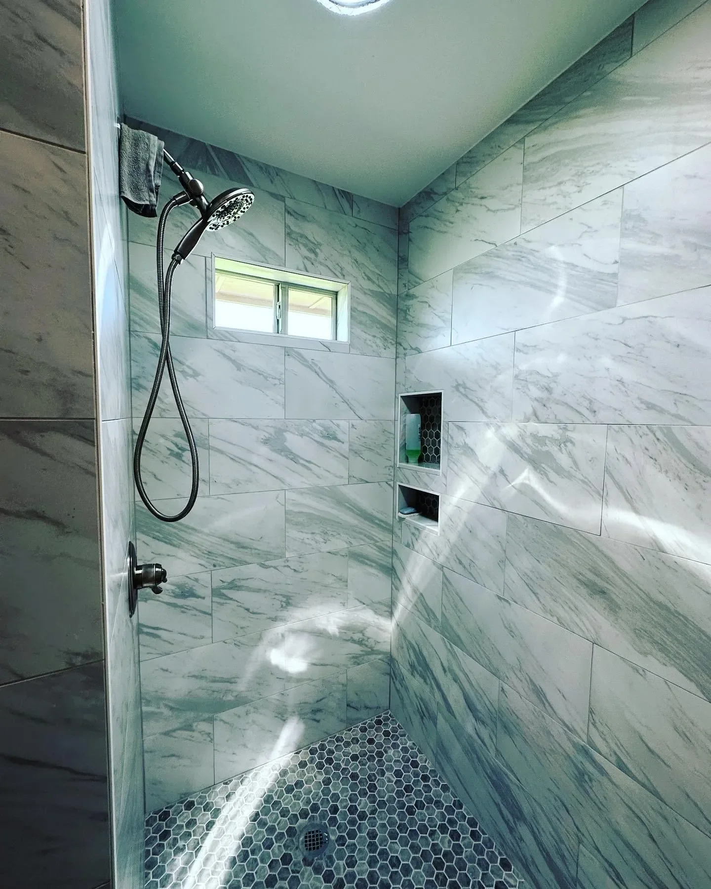 A modern walk-in shower with marble tiles, a small window, a handheld showerhead, and two built-in niches with toiletries.