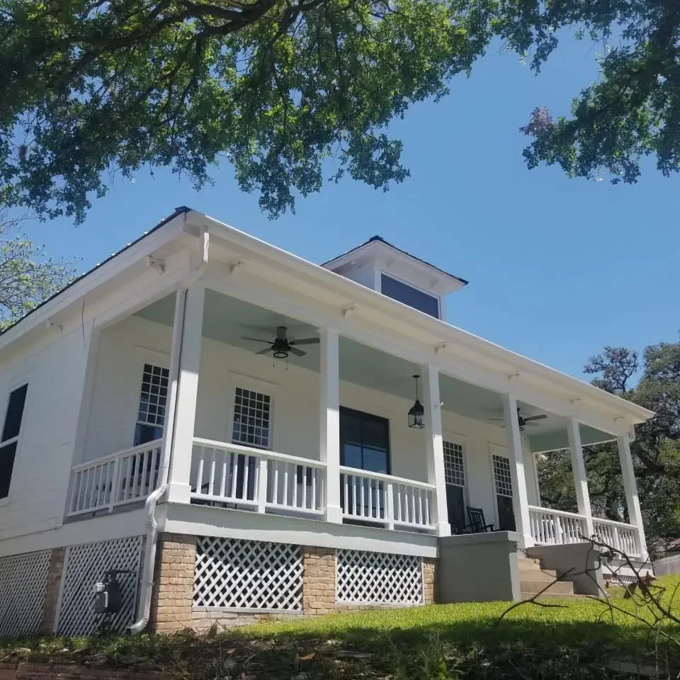 White two-story house with a front porch, black ceiling fans, and hanging lanterns, surrounded by green trees and grass under a clear blue sky.