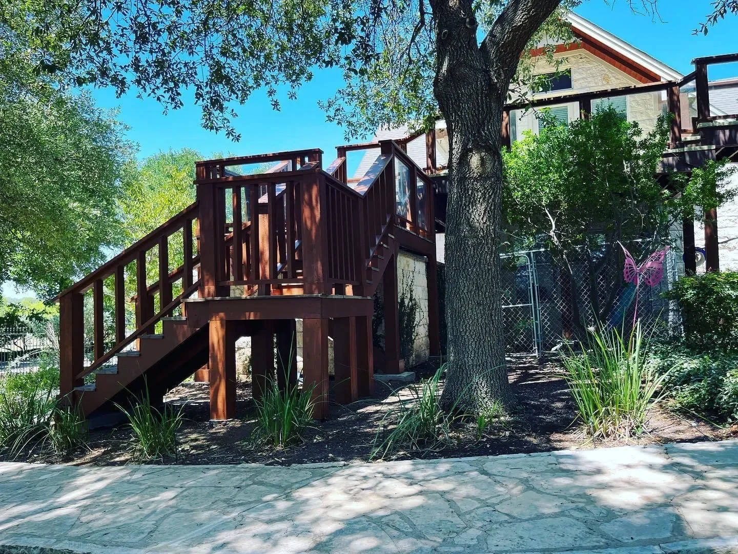 Wooden deck and stairs surrounded by trees, plants, and a house with stone and siding exterior under a clear blue sky.