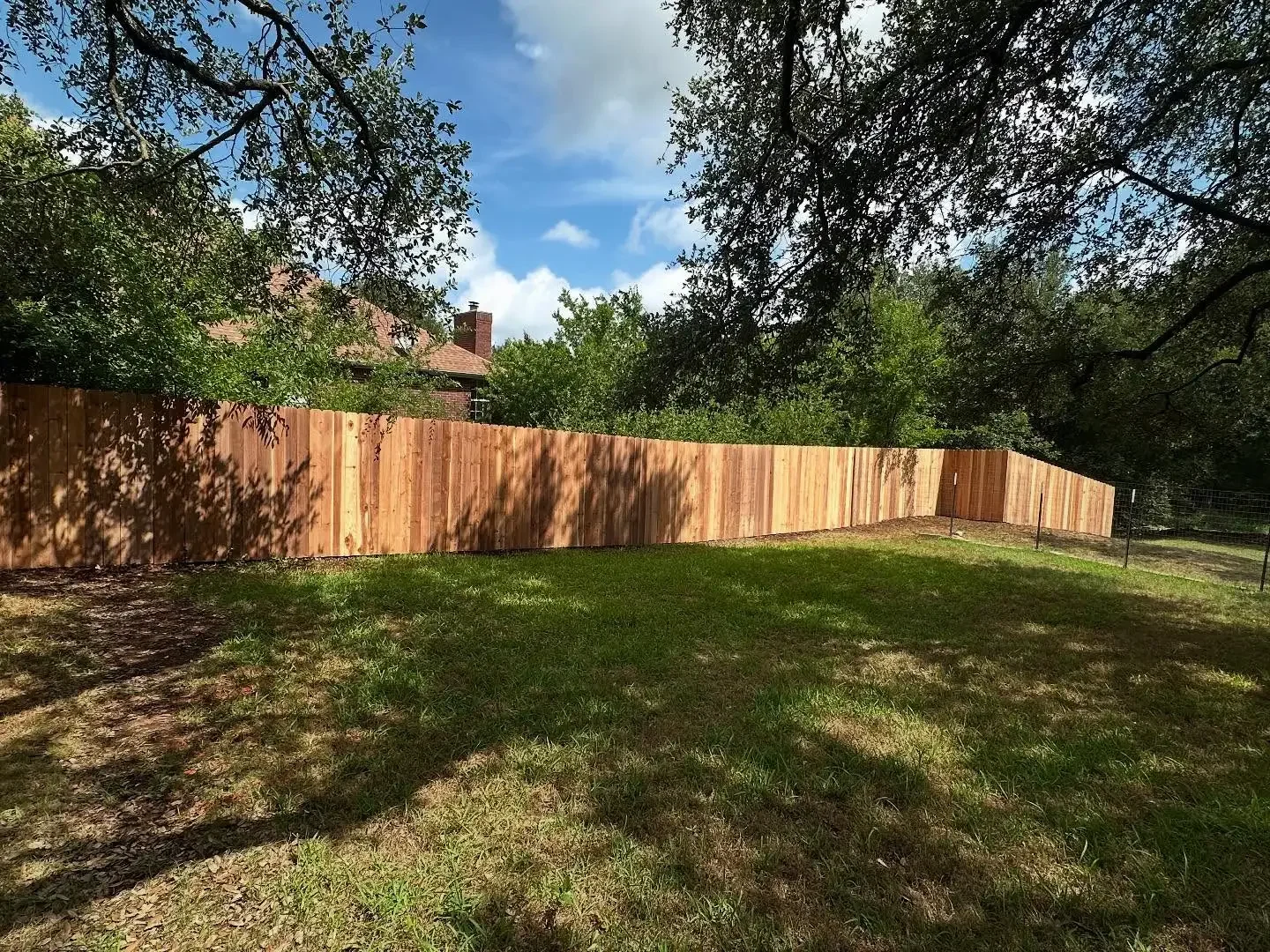 A backyard with a wooden fence, green grass, trees providing shade, and a house with a chimney in the background under a partly cloudy sky.