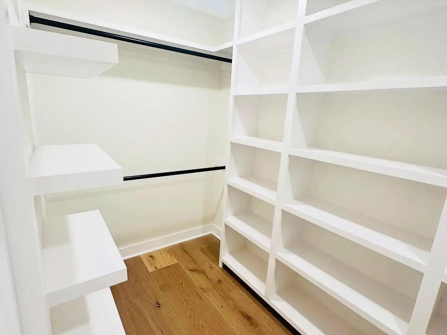 Empty white walk-in closet with shelves on the right and black rods for hanging clothes on the left, with a wooden floor.