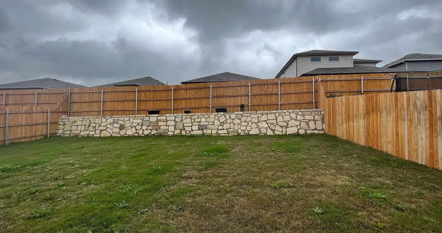 A backyard with a grassy lawn, a stone retaining wall, and a wooden privacy fence. There are three small pots on the wall and gray storm clouds overhead.