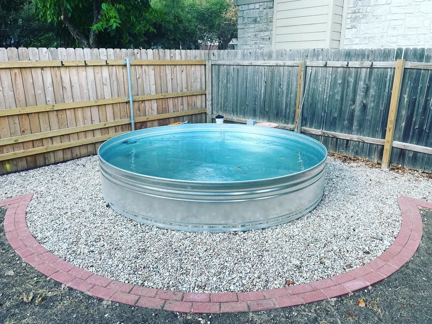 A above-ground metal swimming pool filled with water, situated on a bed of white gravel within a fenced backyard, with trees and a house in the background.