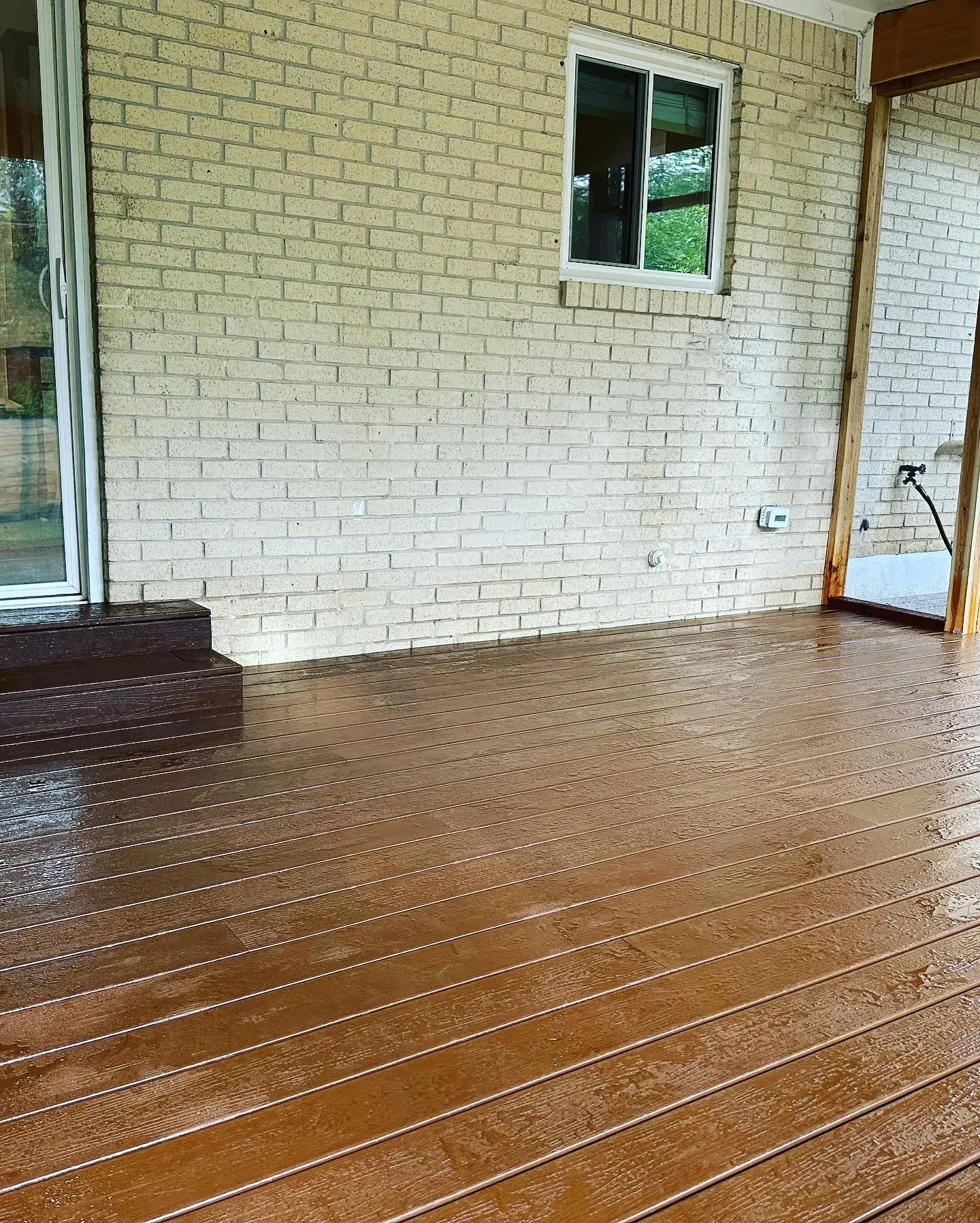 Back porch area with wooden flooring, brick wall, sliding glass door, window, and hose faucet.