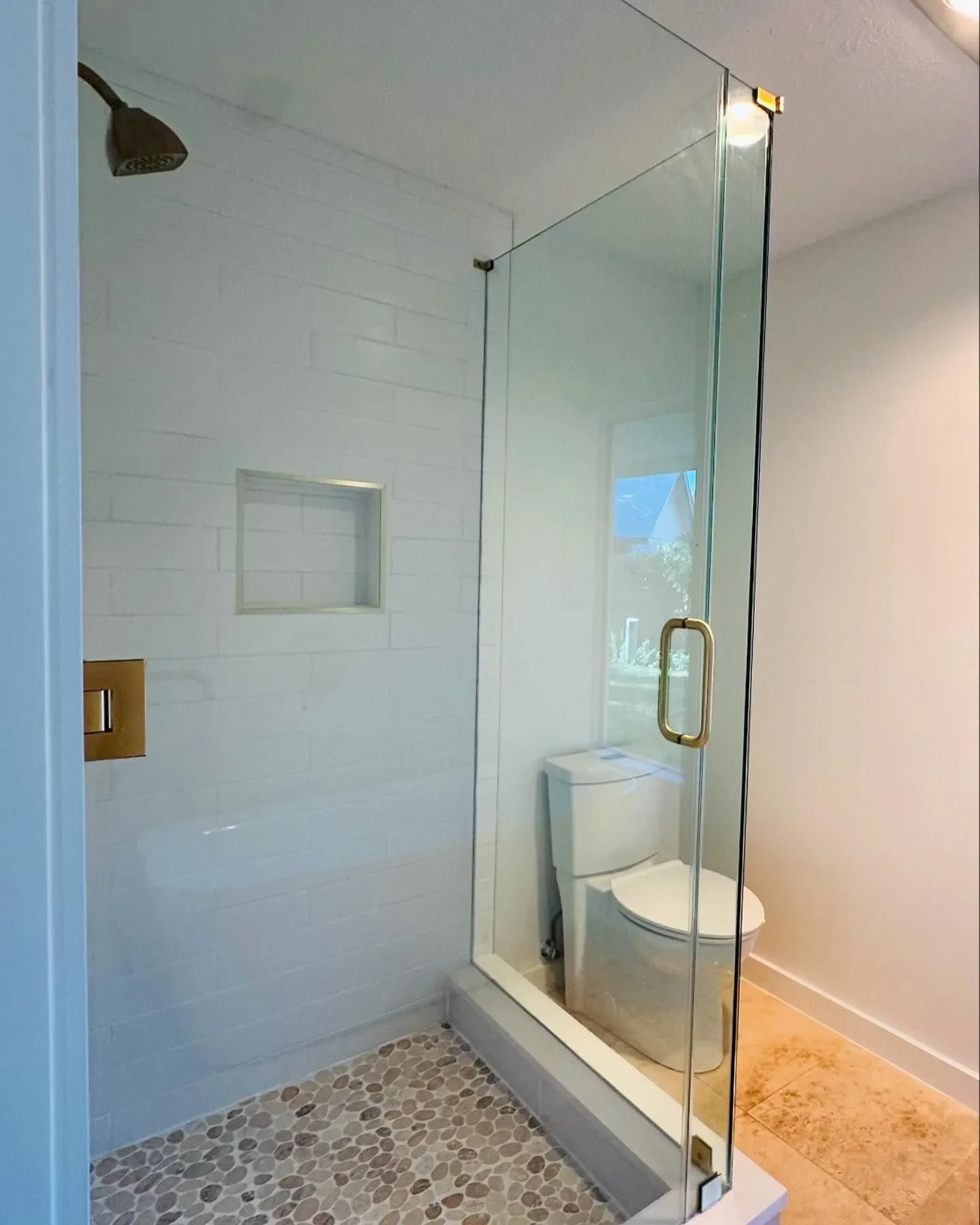 A modern bathroom shower with glass walls and door, a built-in shelf, pebble tile floor, a white toilet, and a window with a view outside.