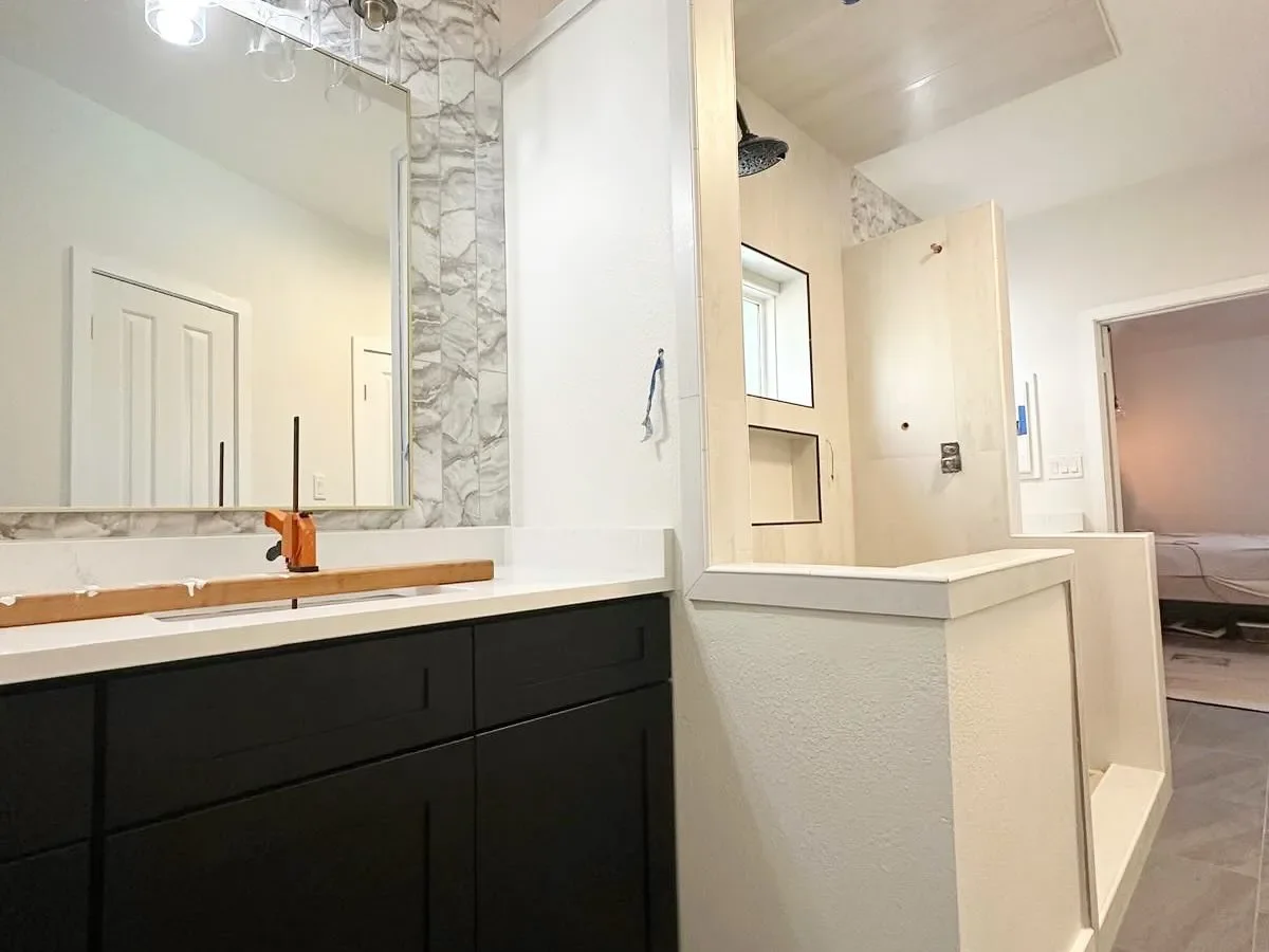 Interior of a bathroom under renovation showing a large mirror, a marble backsplash, black cabinets, and an open shower area with a showerhead, unfinished walls, and a window.