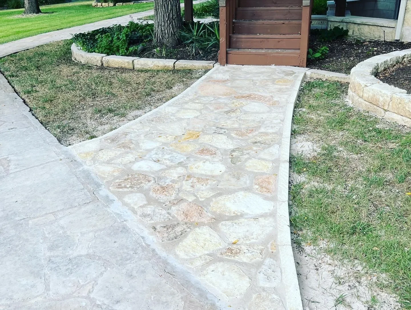 Concrete and stone pathway leading to a wooden gate, with landscaped garden beds on either side and a house in the background.