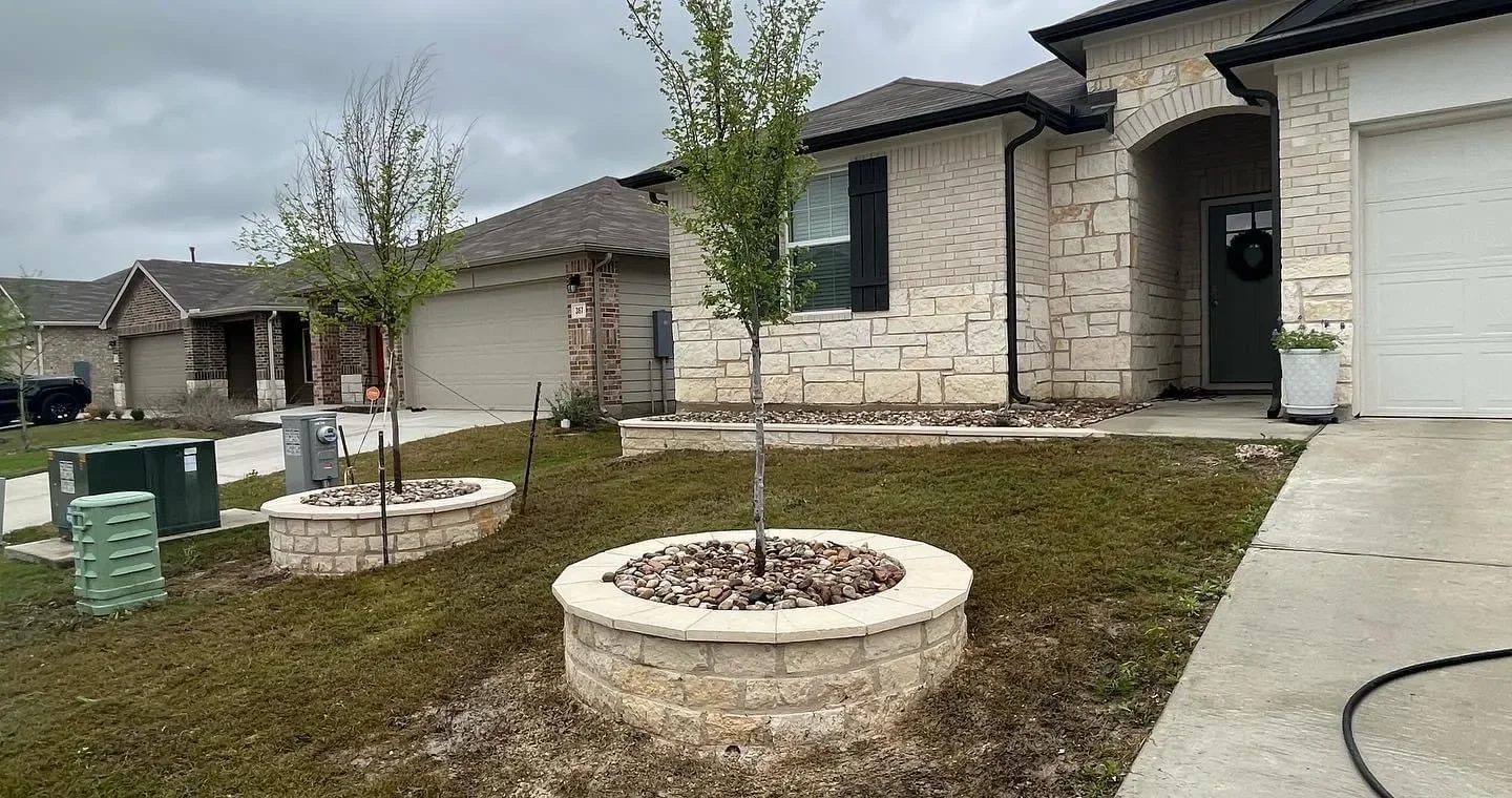 Front yard of a house with small trees planted in circular stone beds, a concrete sidewalk, and a driveway. The house has brick and stone exterior, black shutters, and a wreath on the front door.