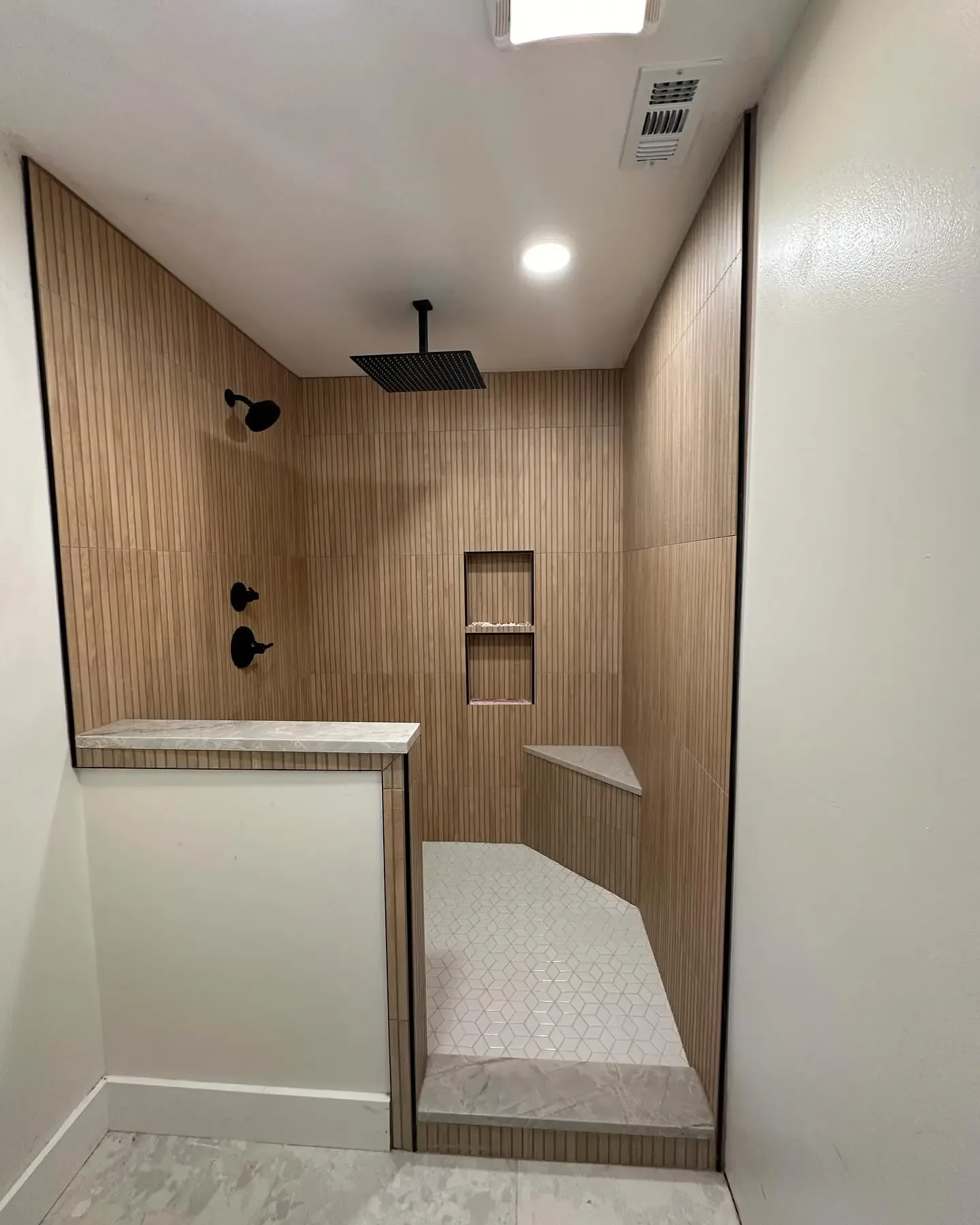 Modern walk-in shower with wood paneling, black fixtures, a large overhead rain showerhead, two smaller wall-mounted showerheads, built-in shelves, and a mixed tile floor.