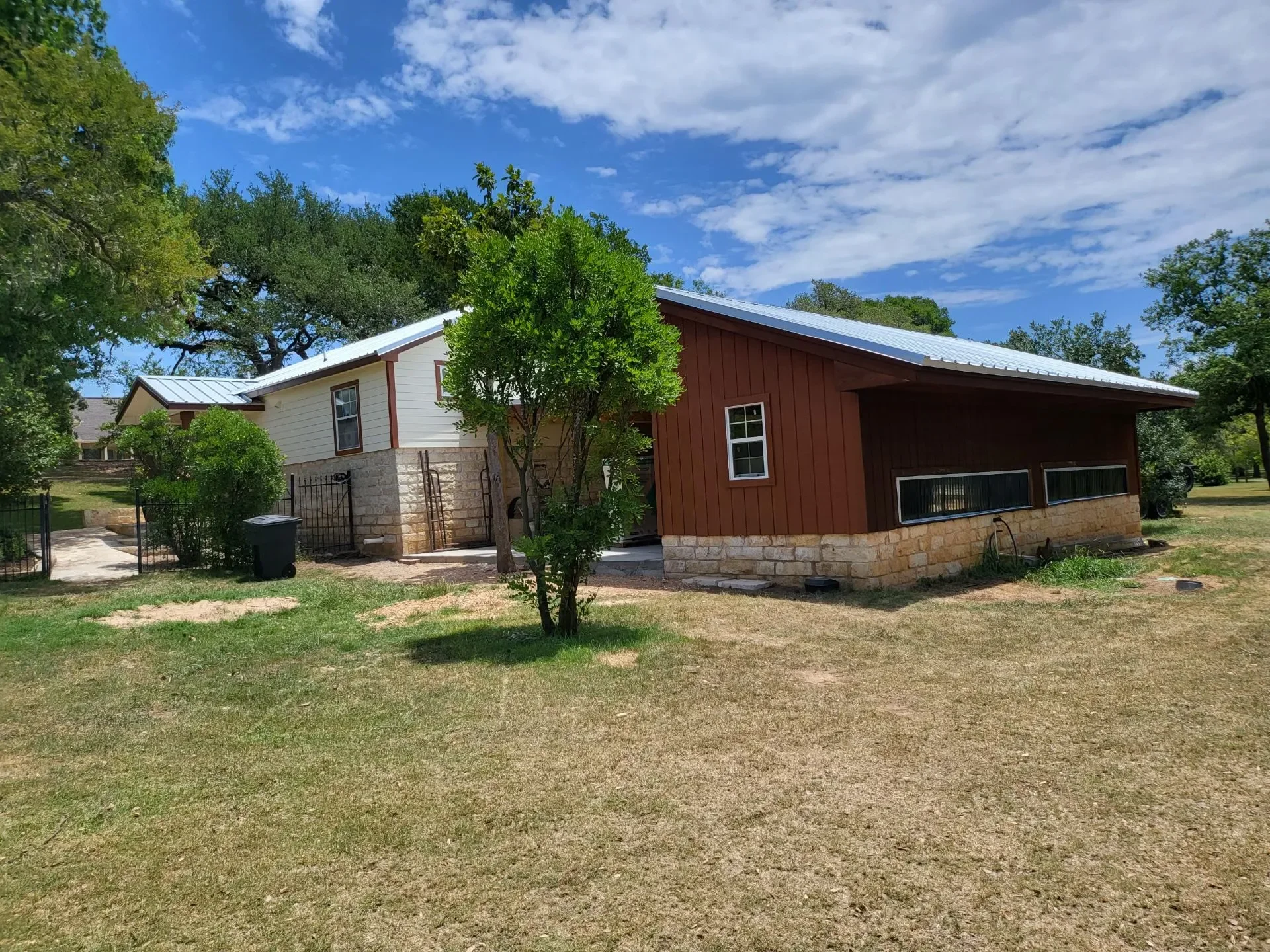 A house with a white upper section and brown wooden siding lower section, stone foundation, metal roof, surrounded by green trees and grass, under a partly cloudy sky.