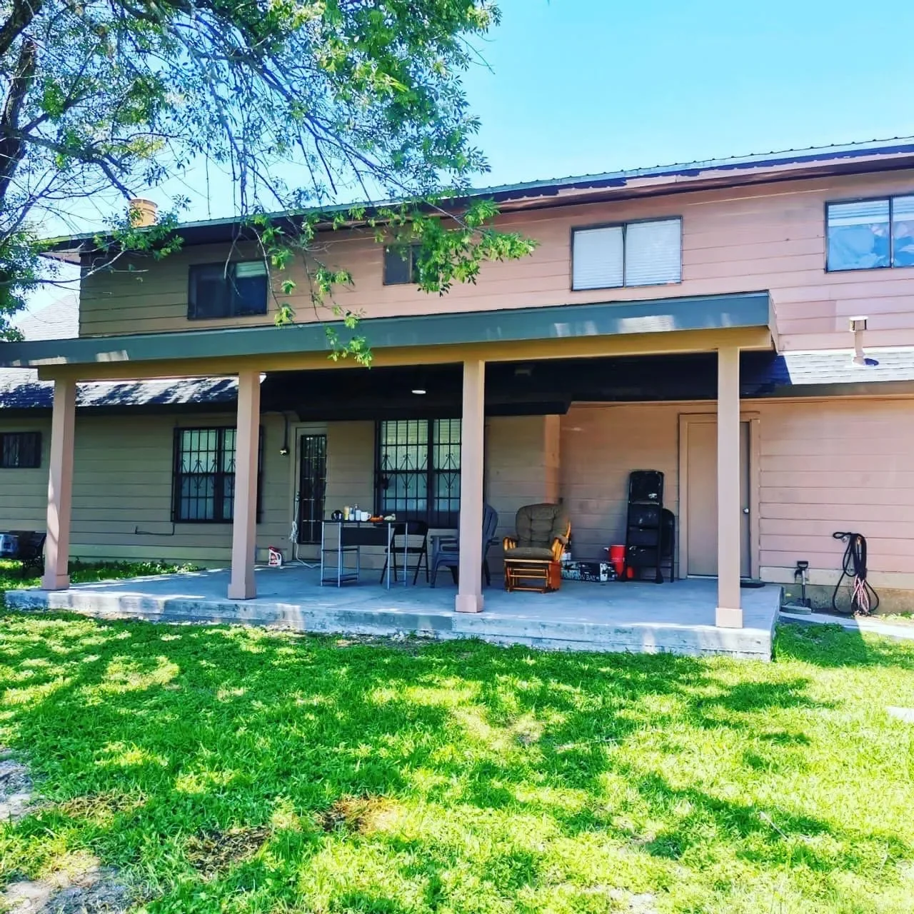Backyard patio with outdoor furniture, chairs, a small table, and a grill, in front of a tan two-story house with windows and a green roof, on a sunny day with green grass and a tree.