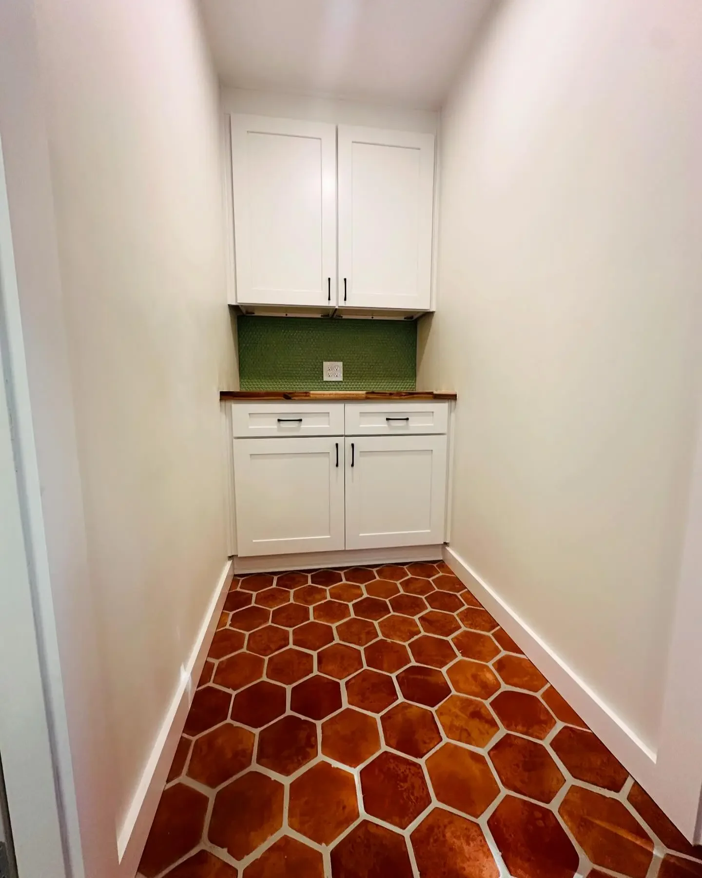 Small kitchen area with white cabinets, green backsplash, wooden countertop, electrical outlets, and brown hexagonal tile flooring.
