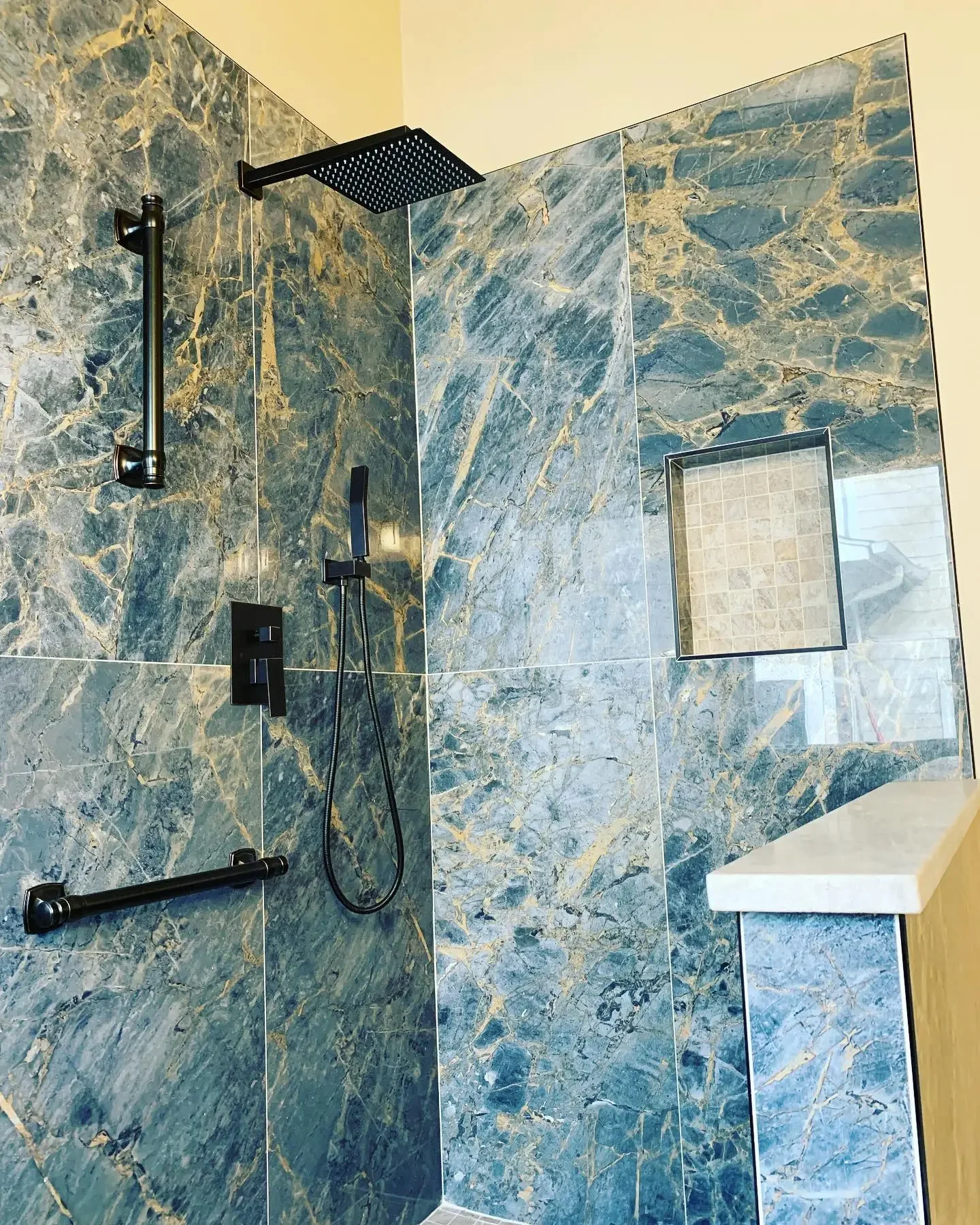 Modern shower with green and gold veined marble tiles, a black rain showerhead, handheld shower, black grab bar, recessed tile niche, and a marble shelf.