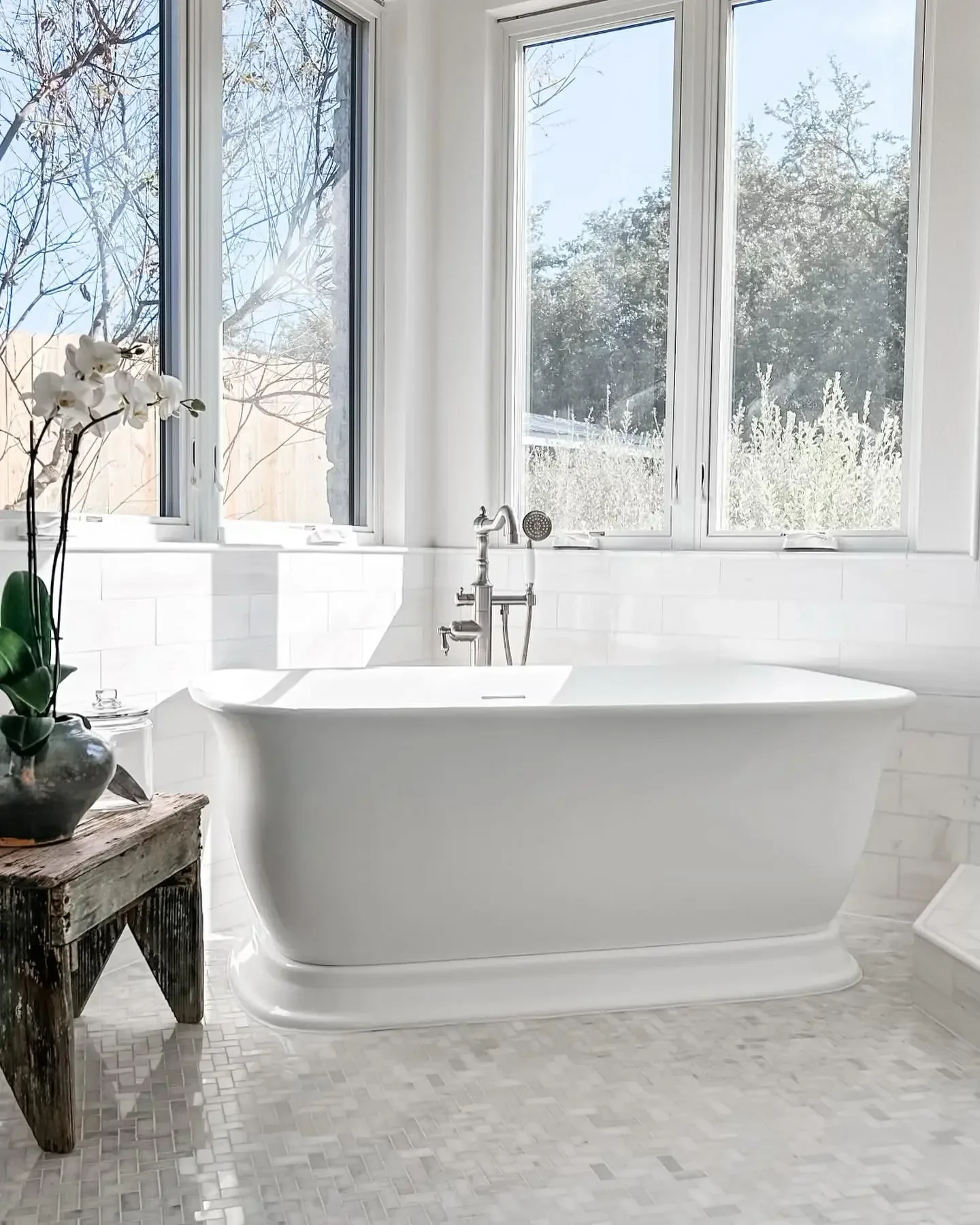A white clawfoot bathtub in a bright bathroom with large windows showing trees outside, a wooden side table with a potted orchid, and a tiled floor.