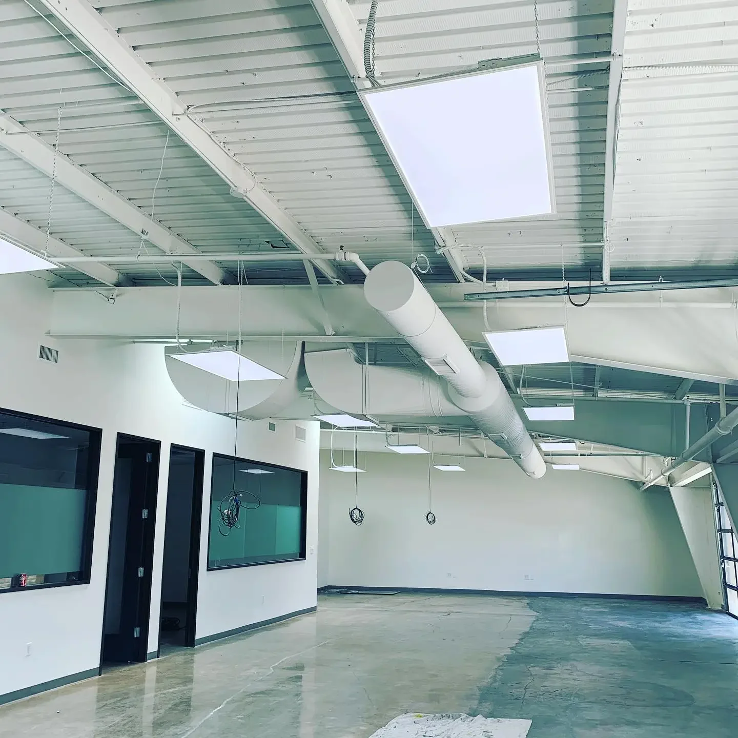 Empty office or commercial space under construction or renovation with exposed ceiling and ductwork, new lighting fixtures, and unfinished flooring.