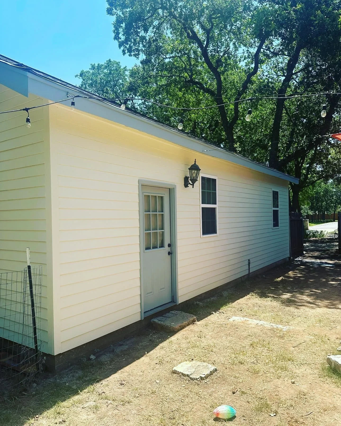 A yellow house with white trim, a door with a window, two windows, an outdoor light, and string lights hanging above. There are trees and a clear blue sky in the background, with a ground of dirt and grass and a colorful ball in the foreground.