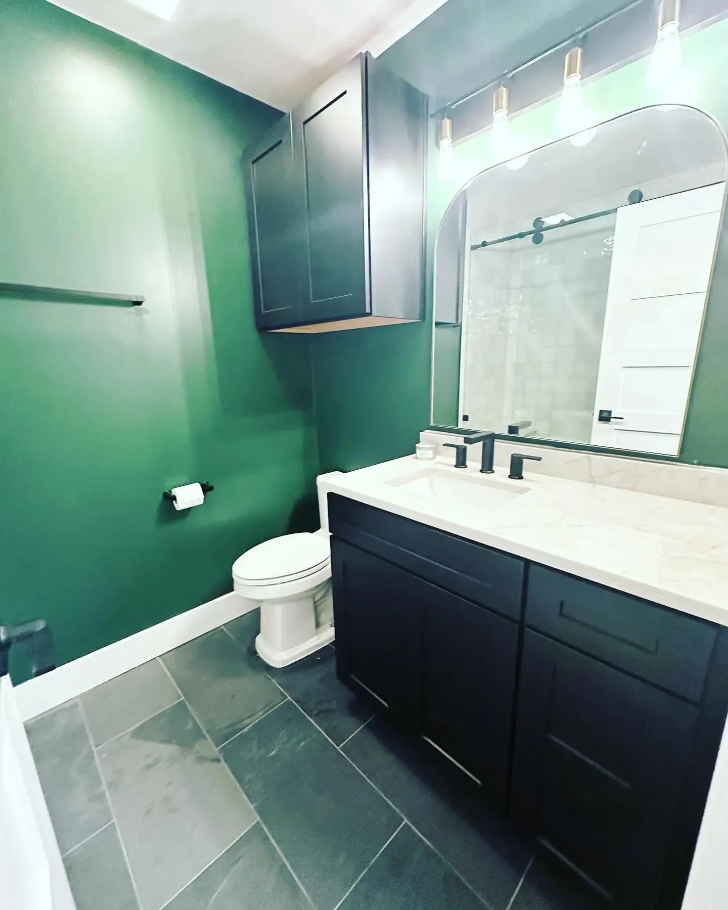 A modern bathroom with dark green walls, a white toilet, a black vanity with a marble countertop, a large mirror, and black fixtures. There is a black cabinet above the toilet and a window with frosted glass letting in natural light.