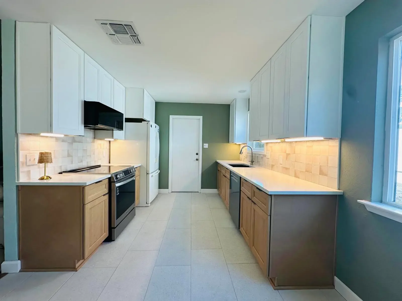 Kitchen with white upper cabinets, light wood lower cabinets, white countertops, beige tile backsplash, stainless steel appliances, green accent wall, beside a window, and with a small lamp on the counter.