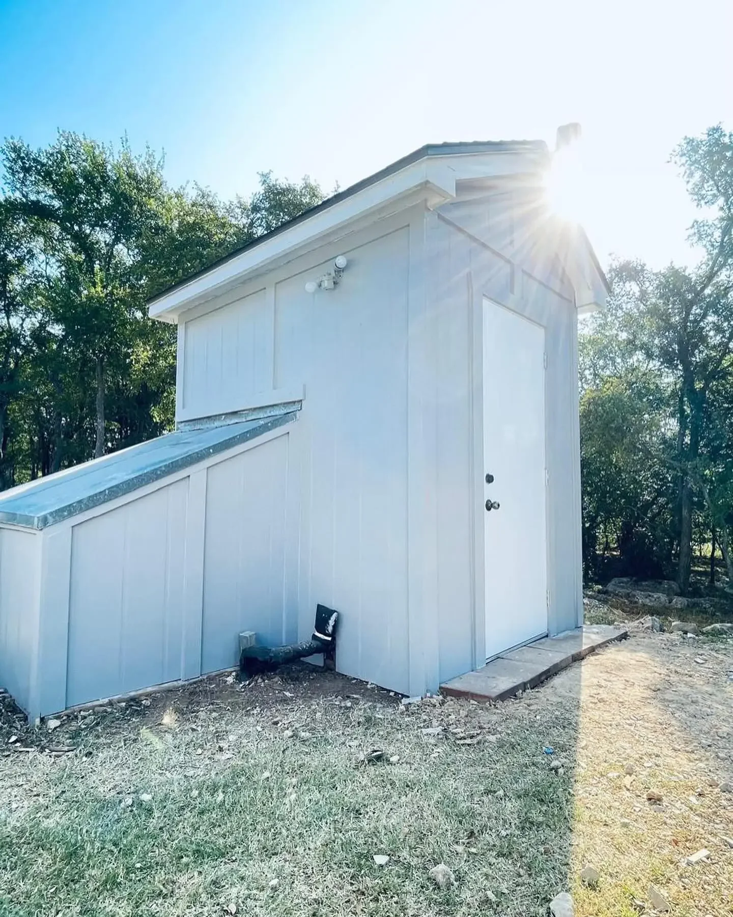 Small white shed with a sloped transparent roof on a grassy area, surrounded by trees, with sunlight shining behind it.