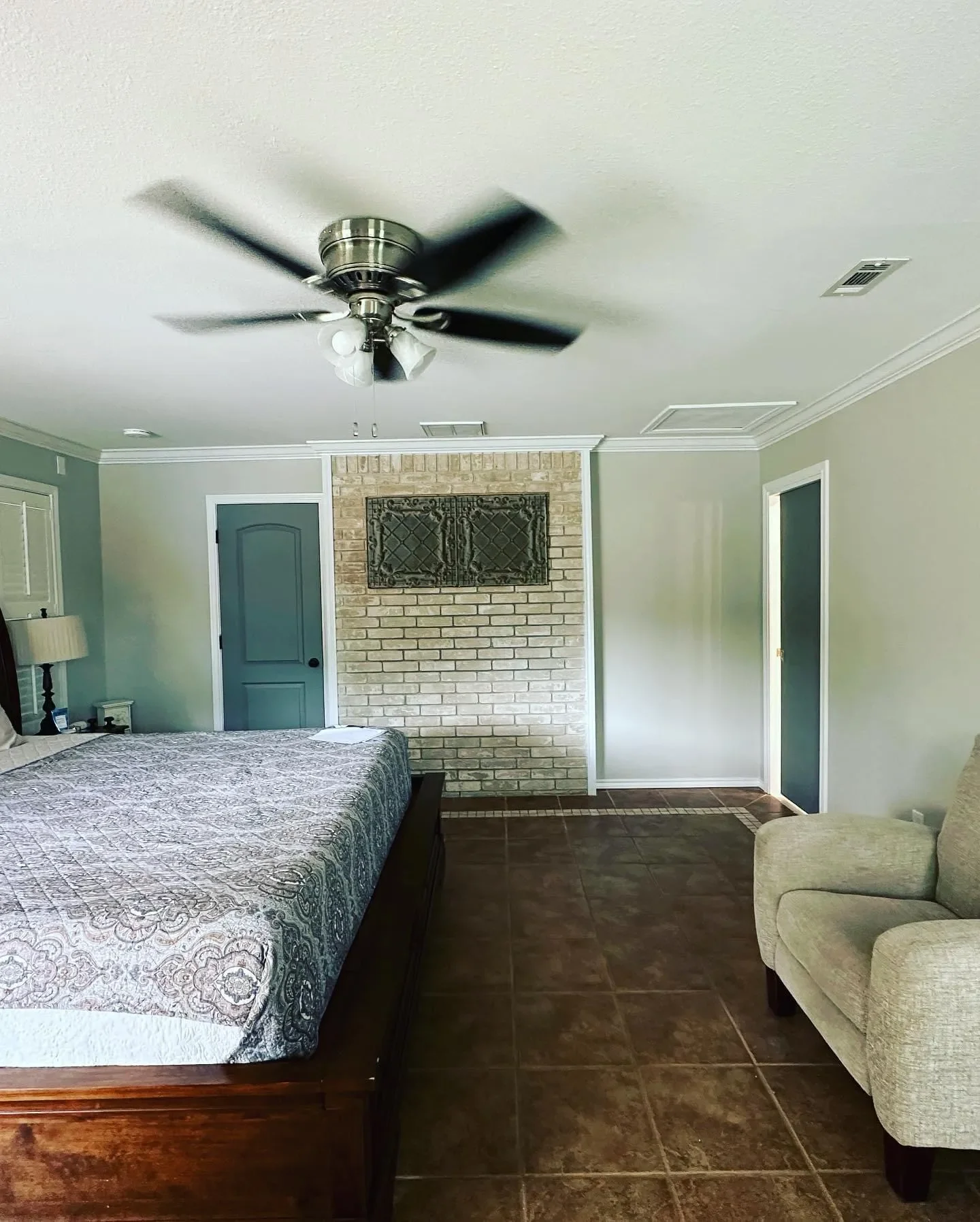 A bedroom with a bed on the left, beige armchair on the right, ceiling fan above, and brick fireplace wall in the background.