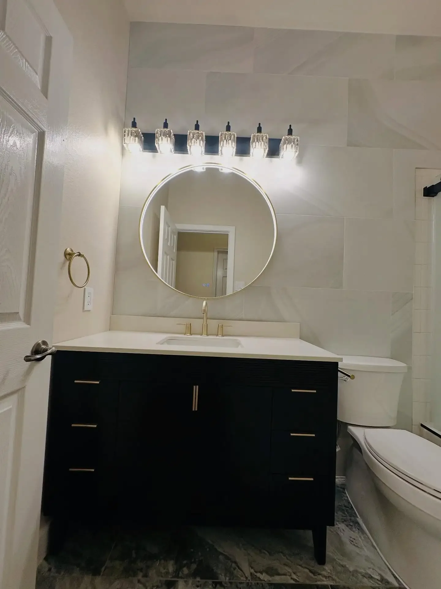 Bathroom with a round mirror, vanity with a black cabinet, white countertop, gold faucet, wall-mounted light fixtures, and a modern toilet.