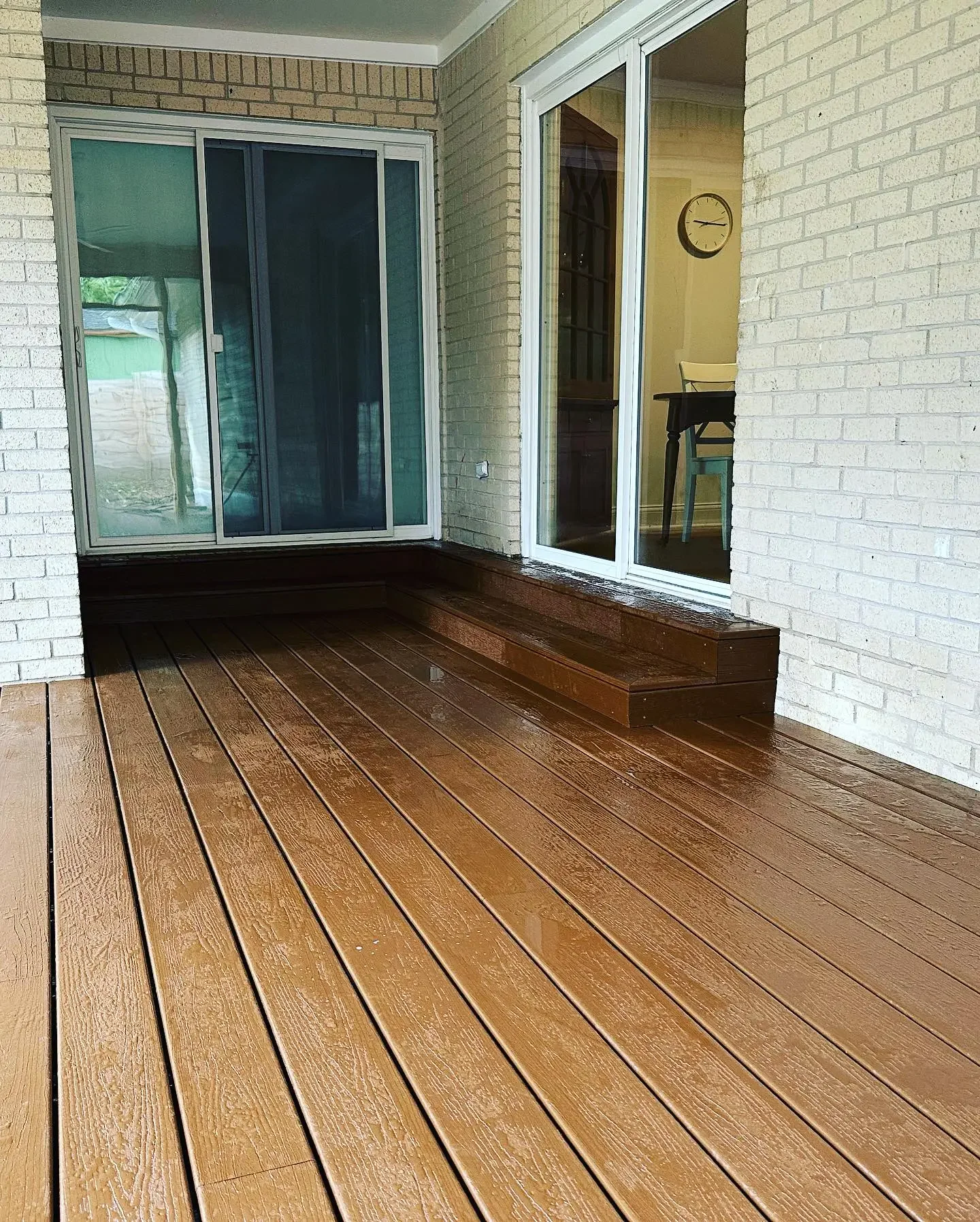 A covered outdoor patio with freshly stained wooden flooring, a step leading up to the sliding glass door, and a brick wall on each side.