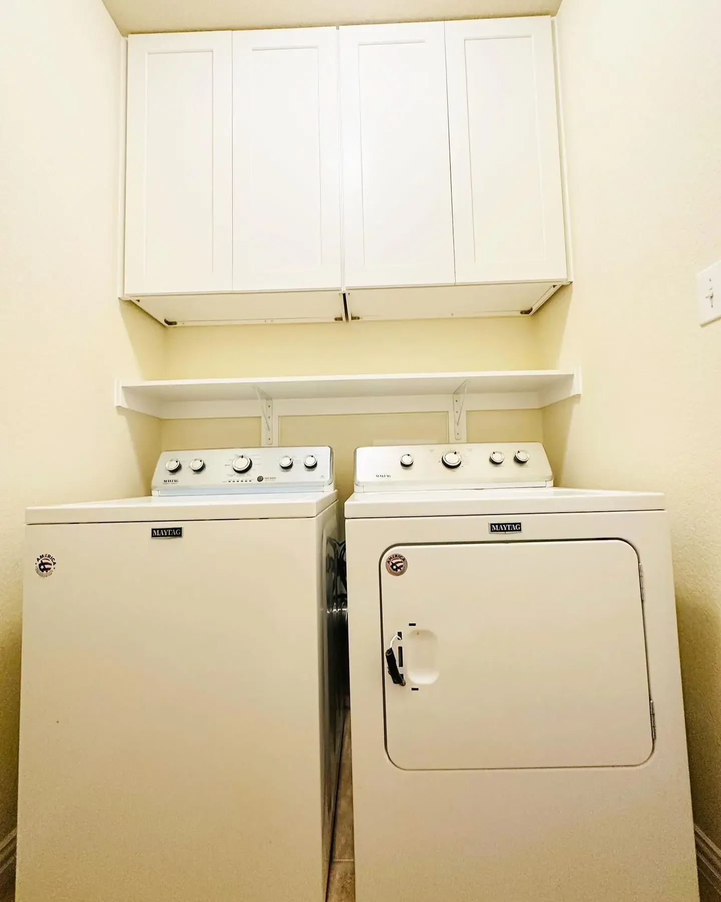 A white top-loading washing machine and a white front-loading dryer in a small laundry room, with white shelves and a cabinet above.