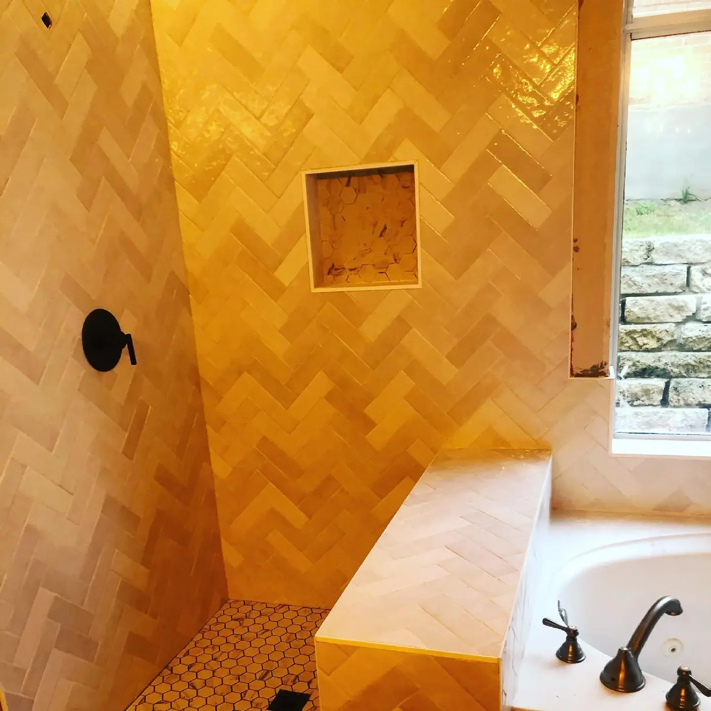 A bathroom shower with yellow wood herringbone wall tiles, a hexagonal tile floor, a small square niche in the wall, a window with stone exterior, and a bathtub with black fixtures.