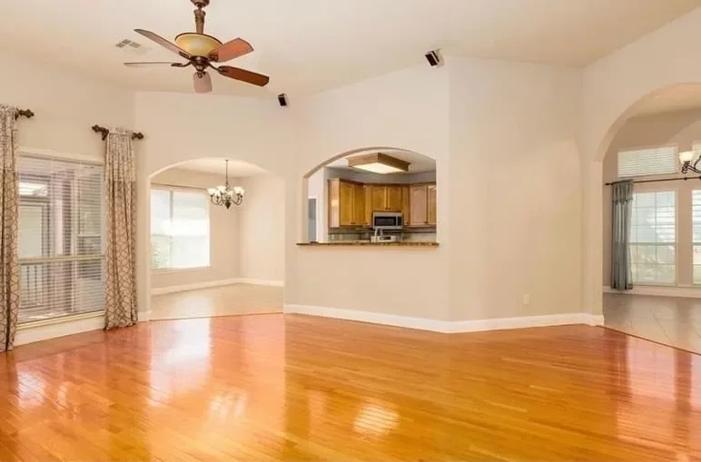 Empty living room with hardwood floors, ceiling fan, windows with curtains, and view into kitchen with wooden cabinets.