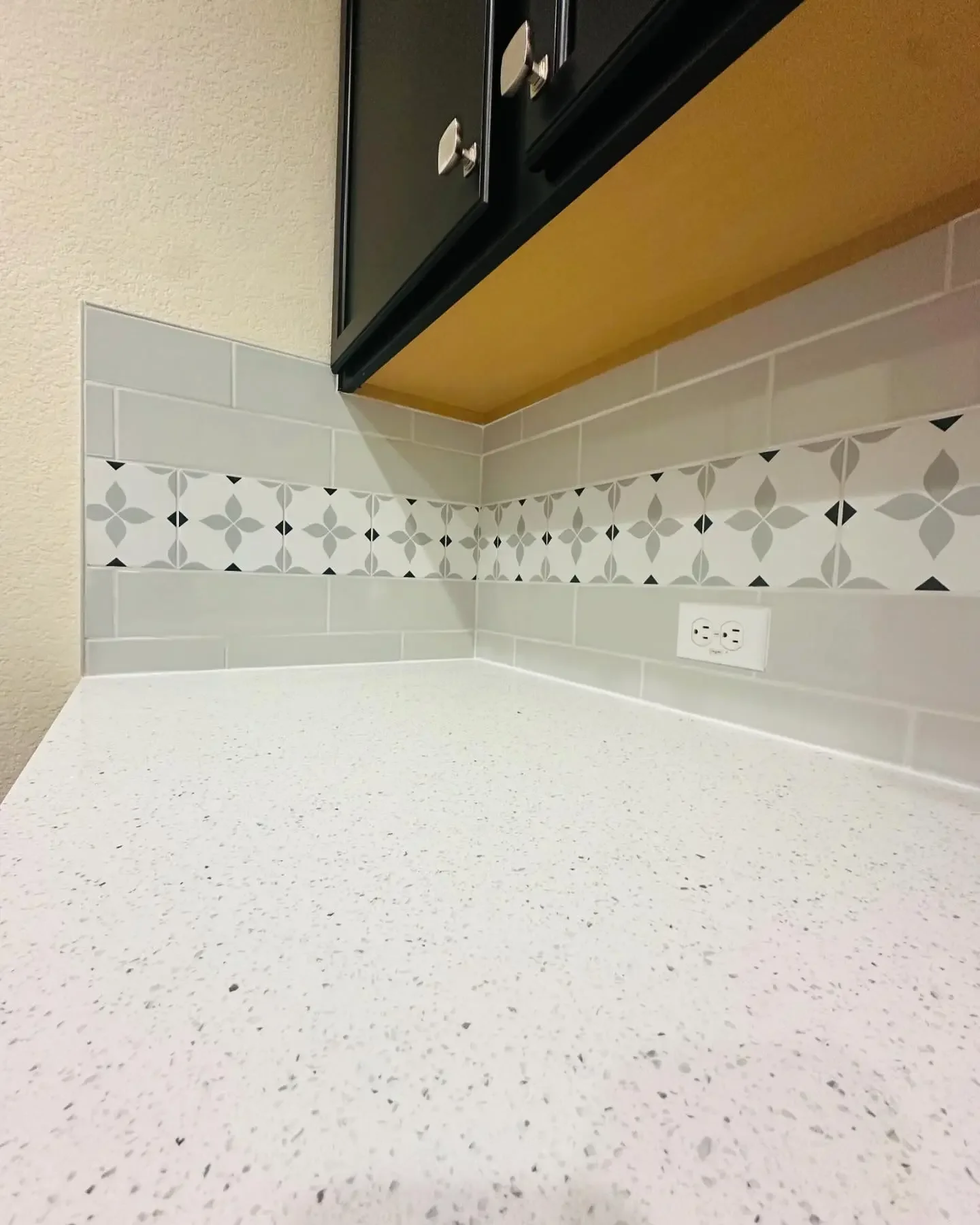 Close-up of a kitchen corner showing a white speckled countertop, a tiled backsplash with gray and patterned tiles, a black upper cabinet, and a beige textured wall.