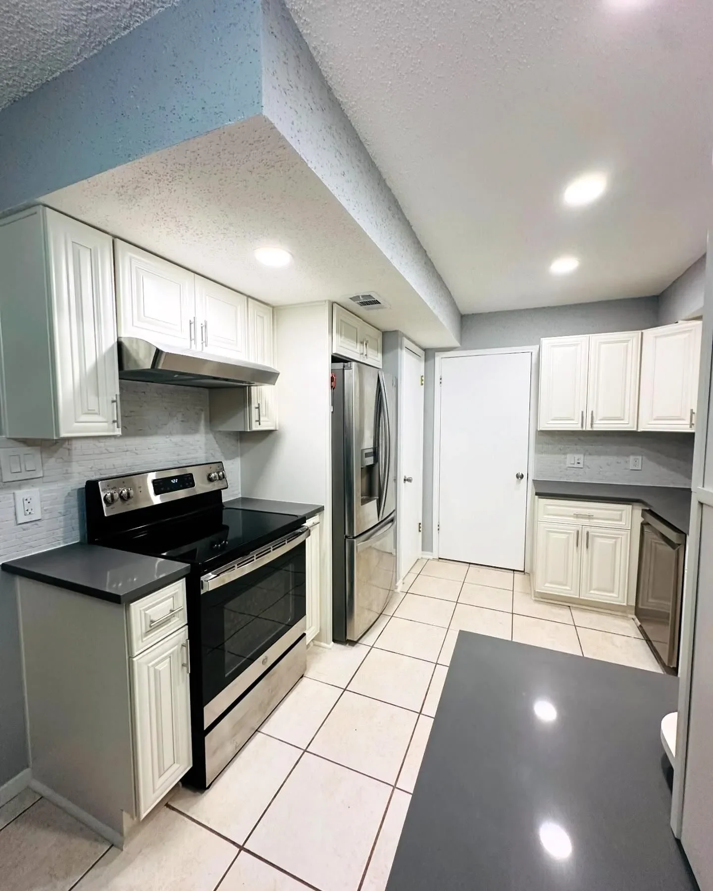 Kitchen with white cabinets, black countertops, stainless steel oven and refrigerator, tile floor, and recessed ceiling lights.