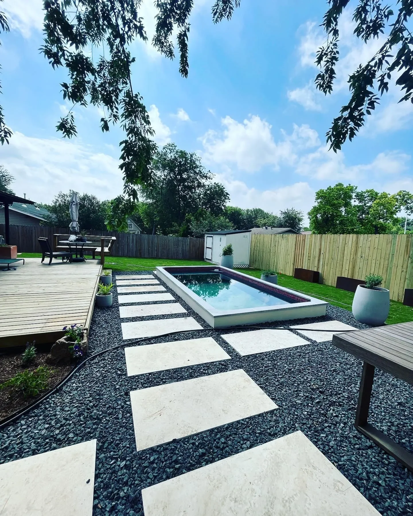A backyard with a small pool, a wooden deck, a paved pathway, potted plants, and outdoor furniture under a blue sky with clouds.