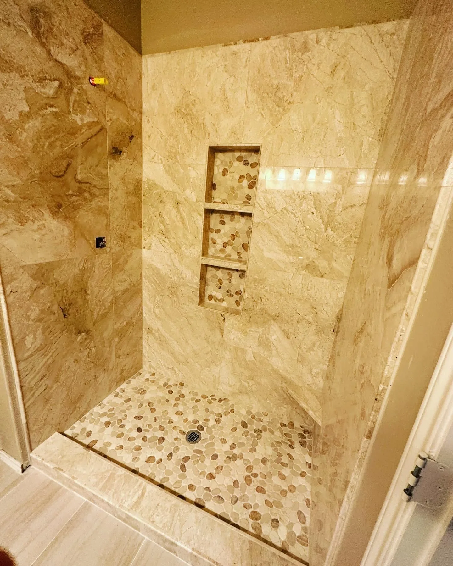 A walk-in shower with beige marble tiles on the walls and a pebble mosaic on the floor. There are three built-in shelves in the wall with a pebble background, and a floor drain in the middle of the shower floor.