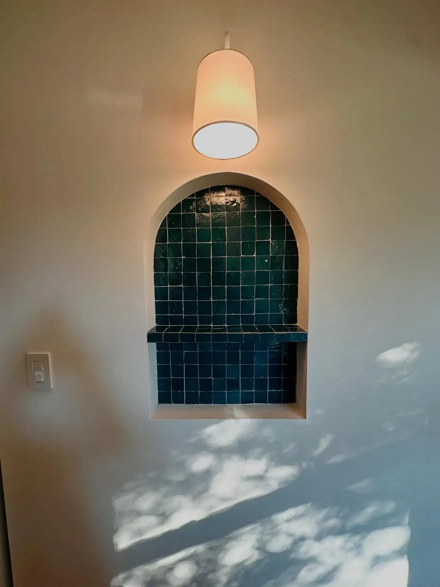 A wall niche with blue tiles and a light fixture hanging above, with sunlight creating a shadow pattern on the wall.