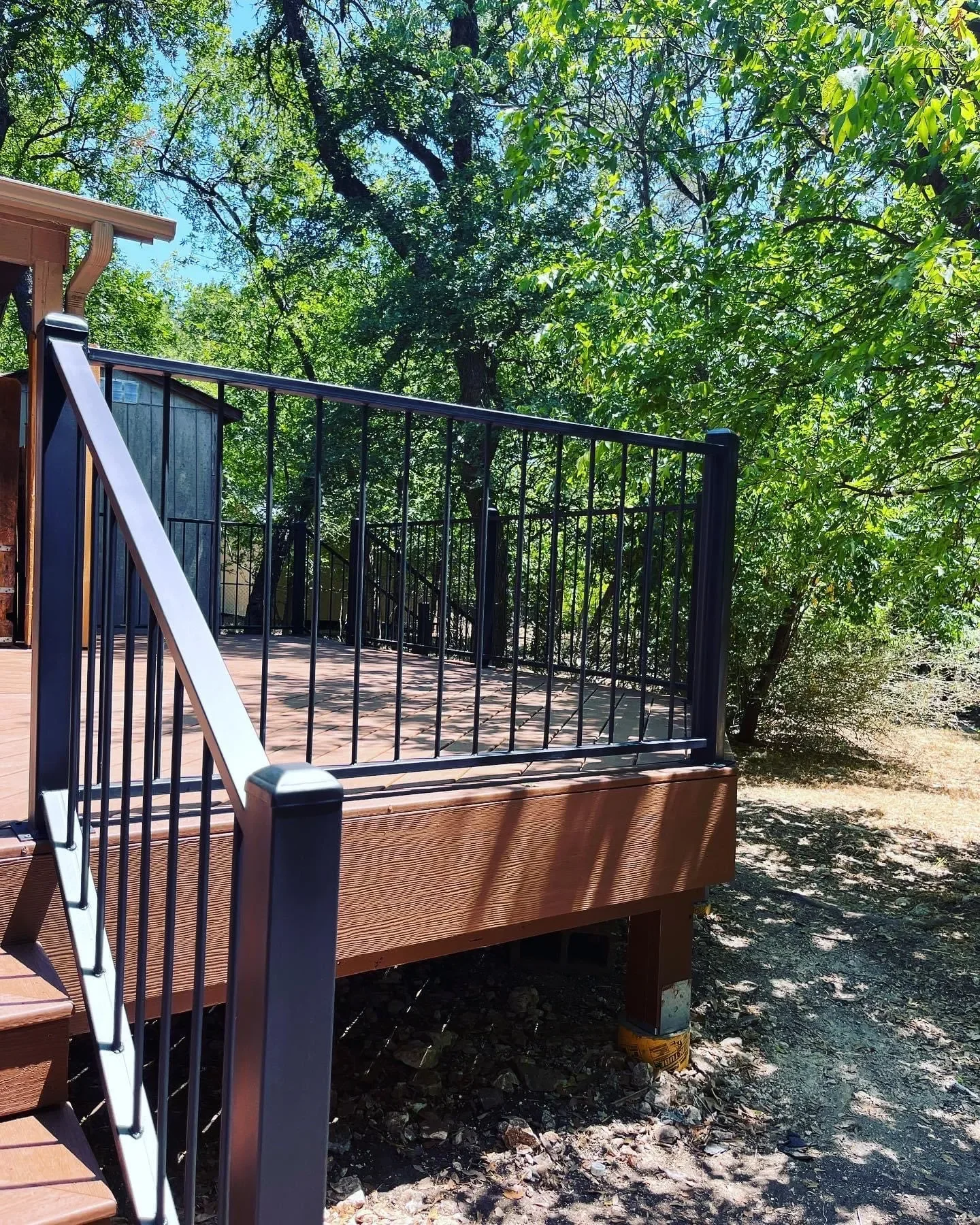 Wooden deck with black metal railing surrounded by green trees and shaded ground.