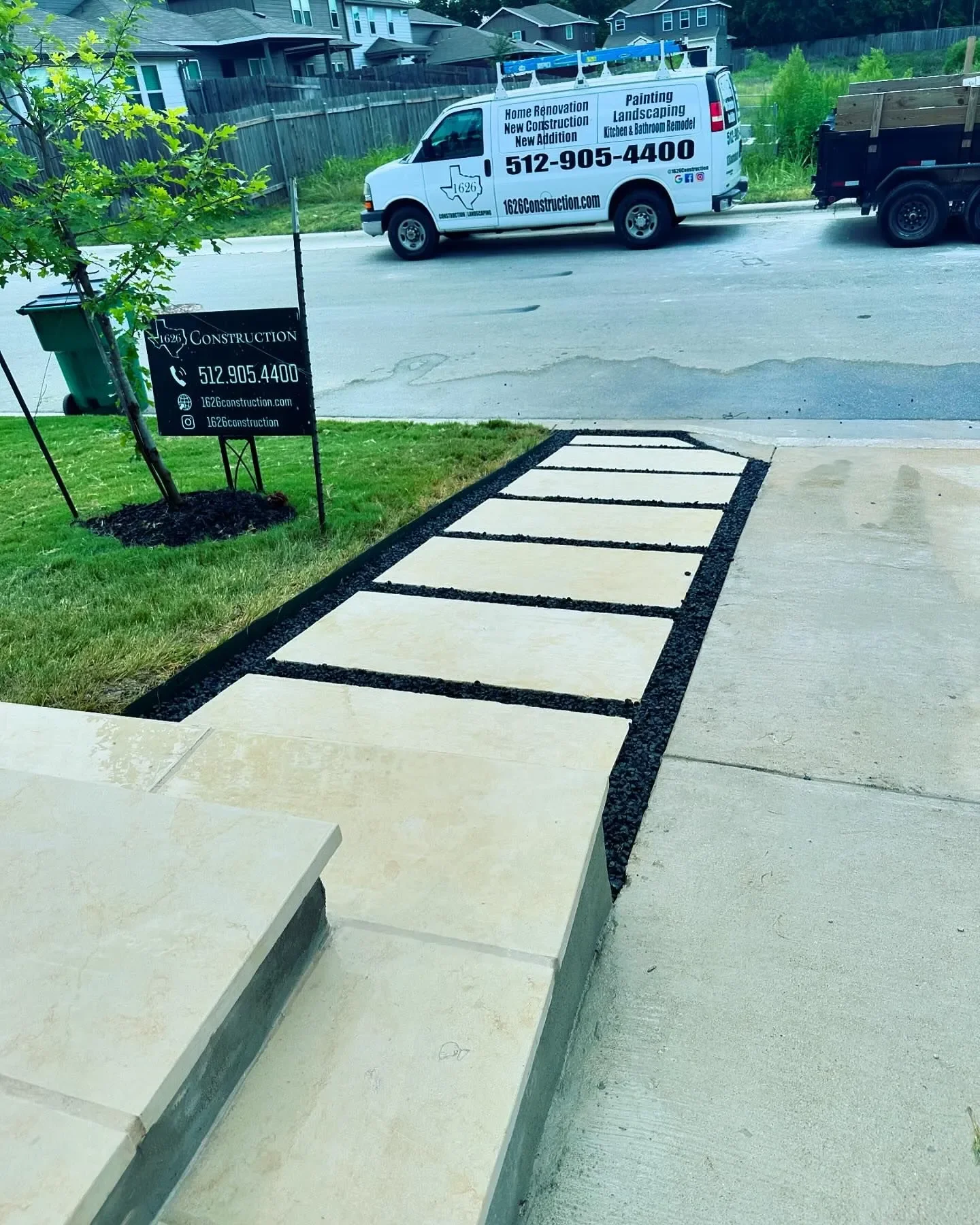 New concrete sidewalk installation with beige tiles bordered by black gravel in front of a house, with a construction sign on the lawn and a construction van on the street.