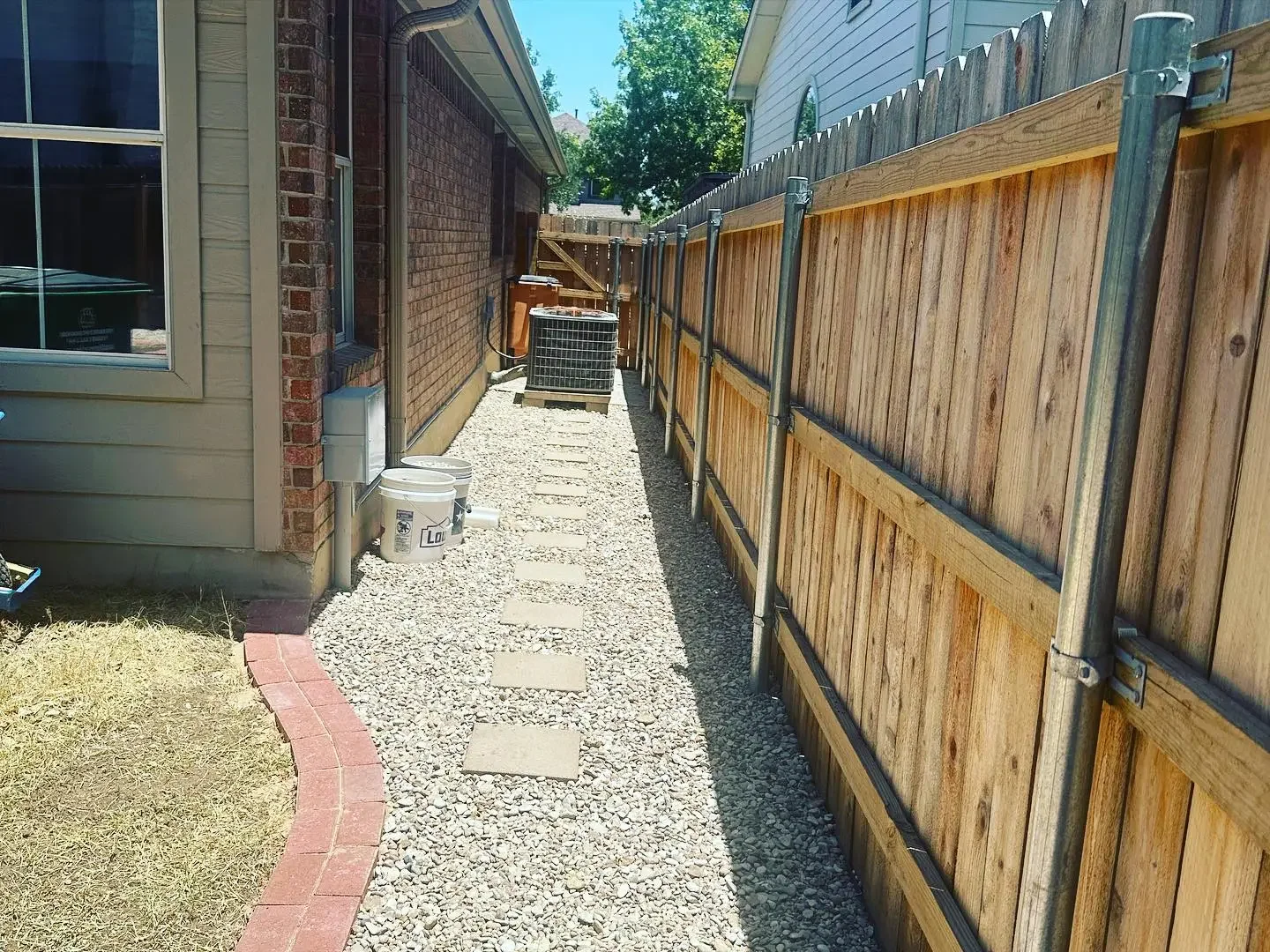 Side yard with gravel pathway, bordered by a brick and grassy lawn on the left, and a wooden fence on the right. The yard includes an air conditioning unit, buckets, and pavers along the pathway, with a house on the left and another house partially v