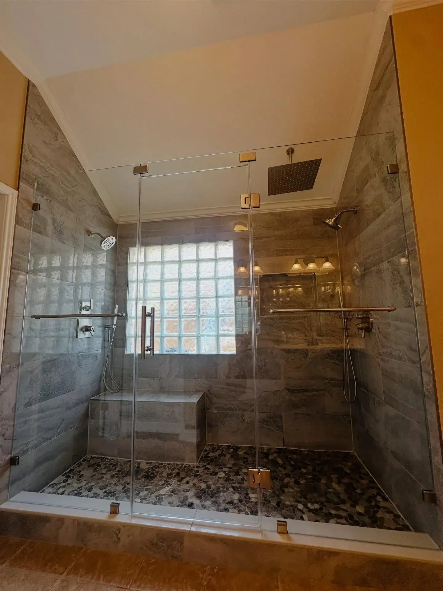 A modern frameless glass shower with two shower heads, one rainfall showerhead on the ceiling and a handheld shower on the wall, a bench with pebble tiles on the floor, and a glass block window letting in natural light.