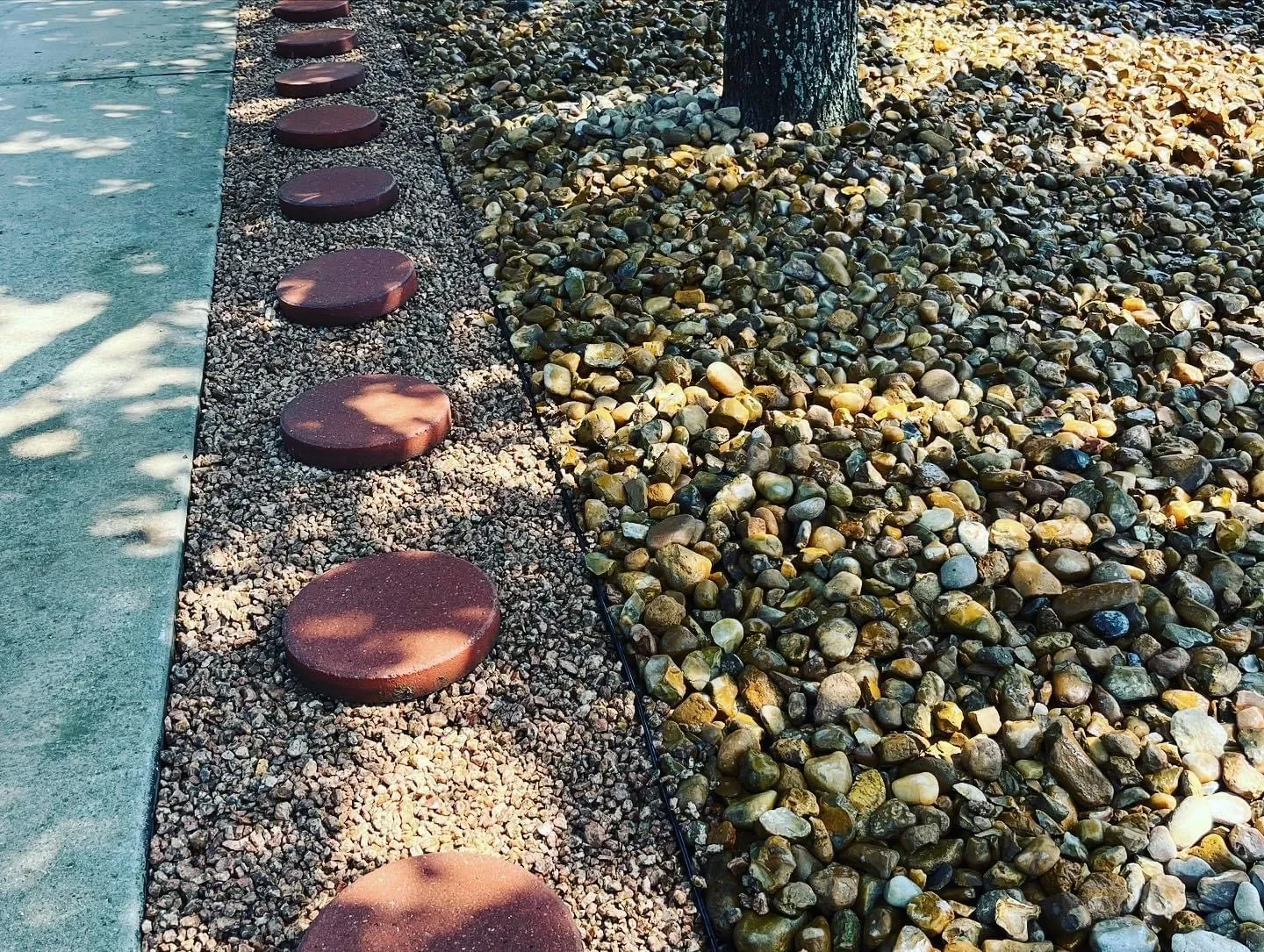 Pathway with large round stepping stones on a bed of small gravel, adjacent to a mulched area with rocks and a tree trunk.