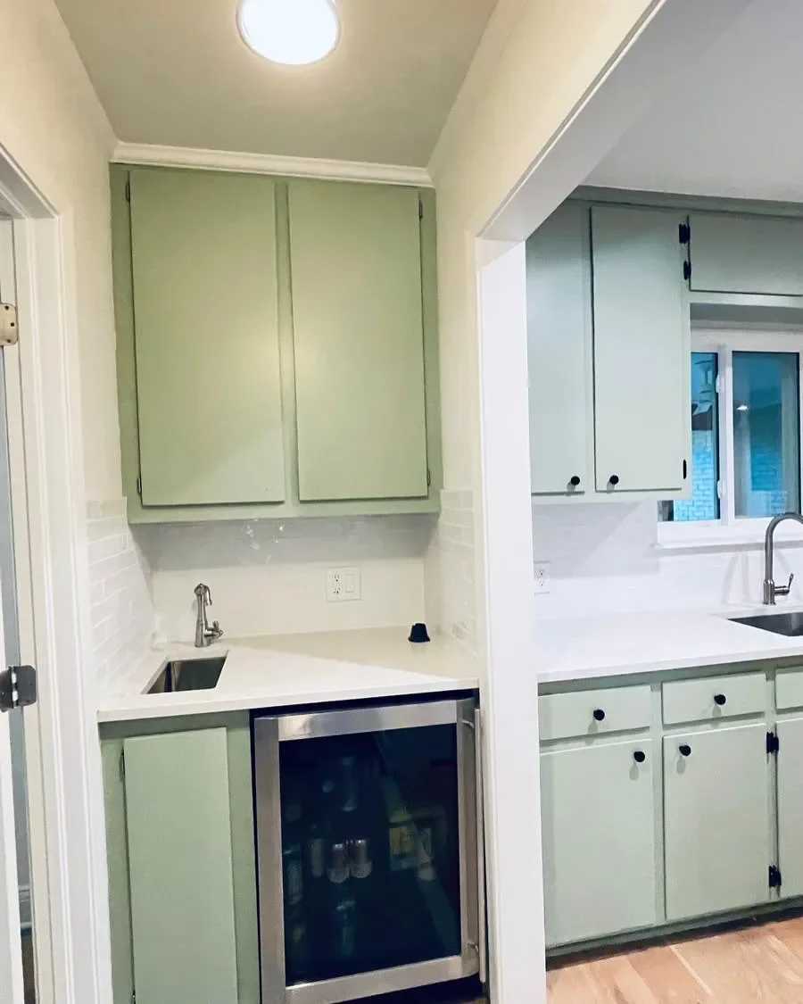 A small kitchenette with pastel green cabinets, a mini fridge with a glass door, a small sink, and a window.