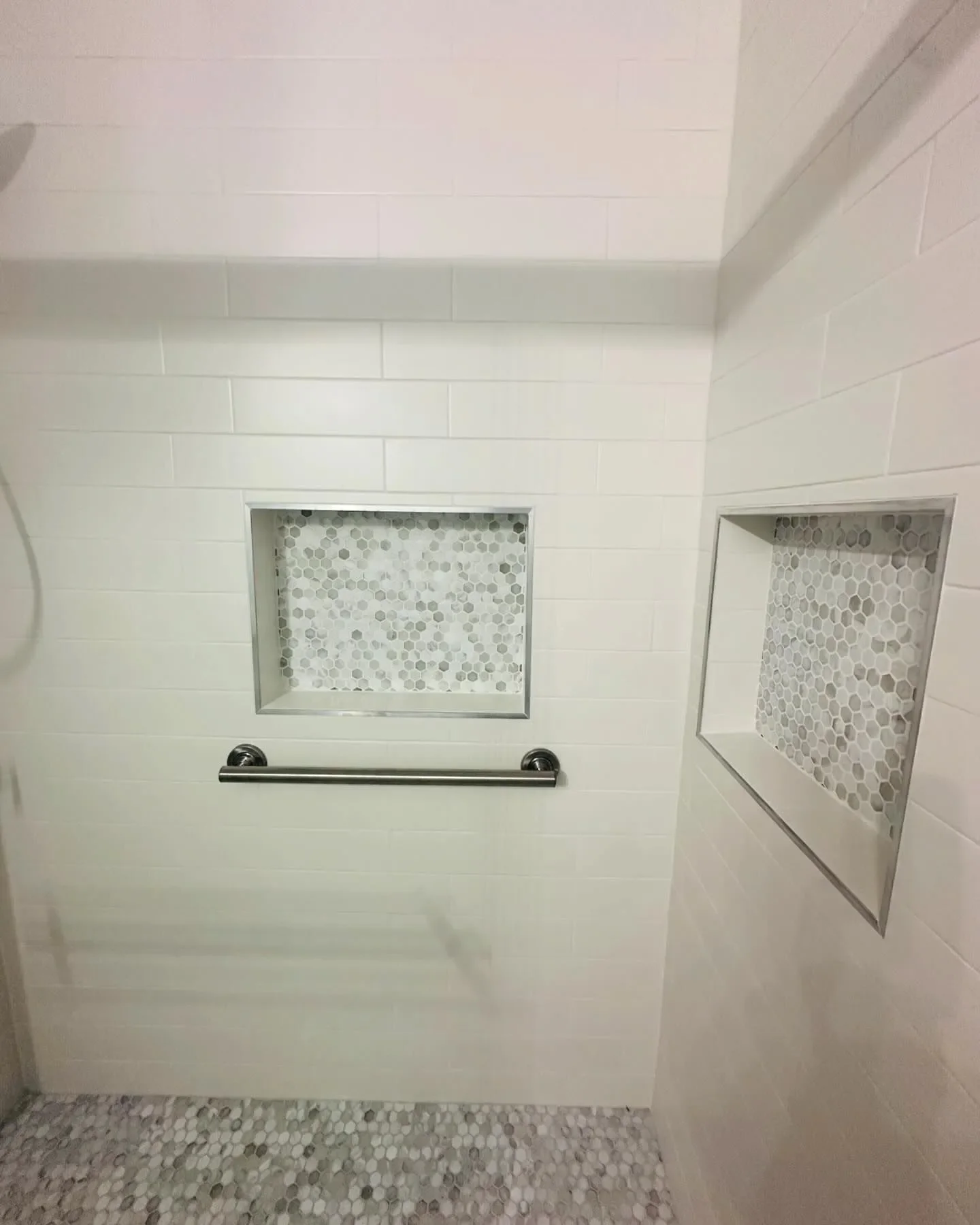 Shower area with white subway tiles, two recessed shower niches with hexagonal mosaic tiles, a metal grab bar, and a pebble tile floor.