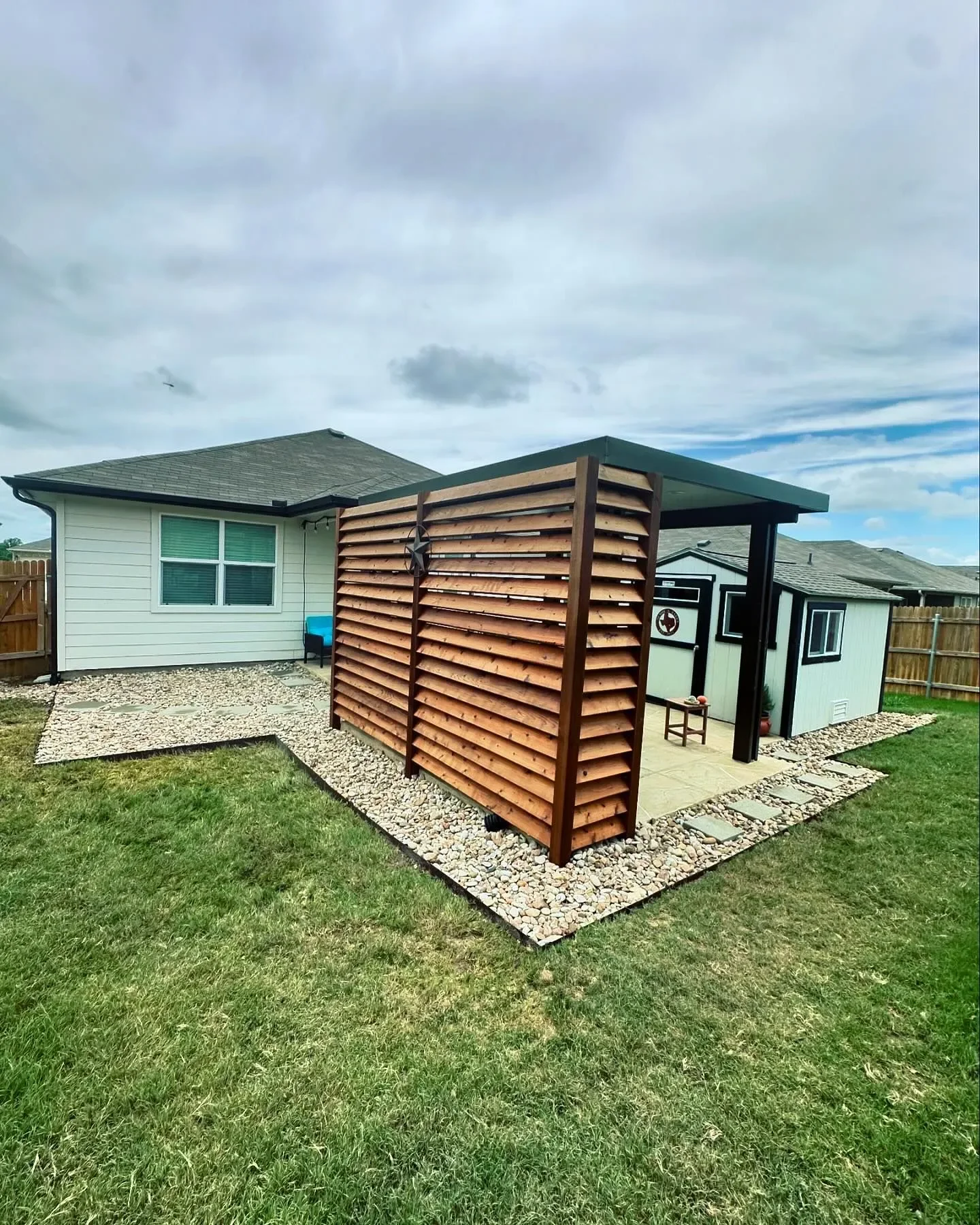 Backyard with a small white house, a shed, and a wooden privacy screen, with gravel pathways and green grass under a cloudy sky.