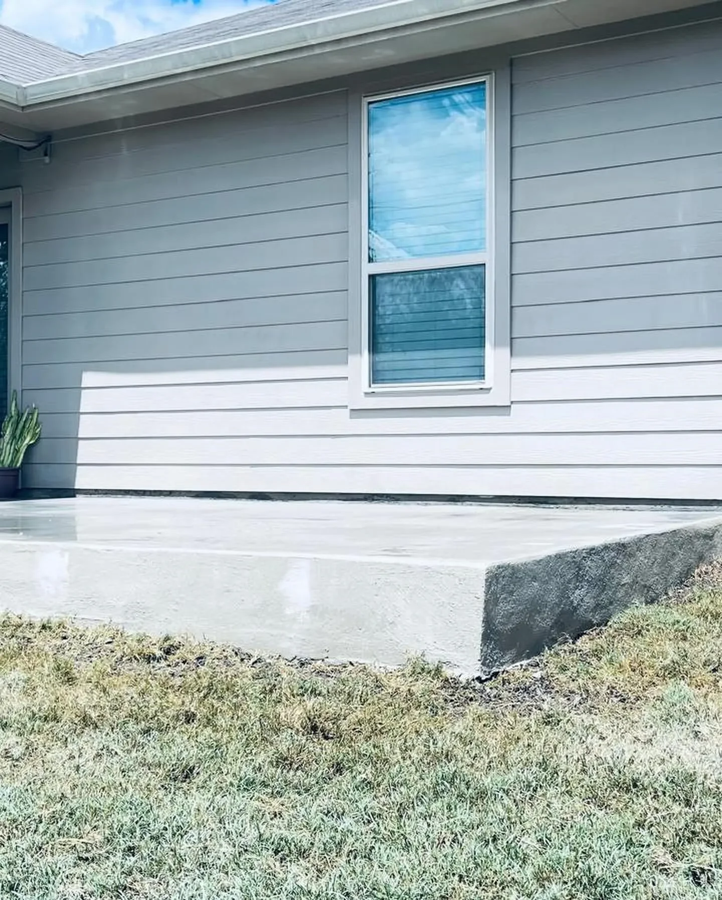 Exterior view of a house with gray siding, a large window, a concrete patio, and a small patch of grass.