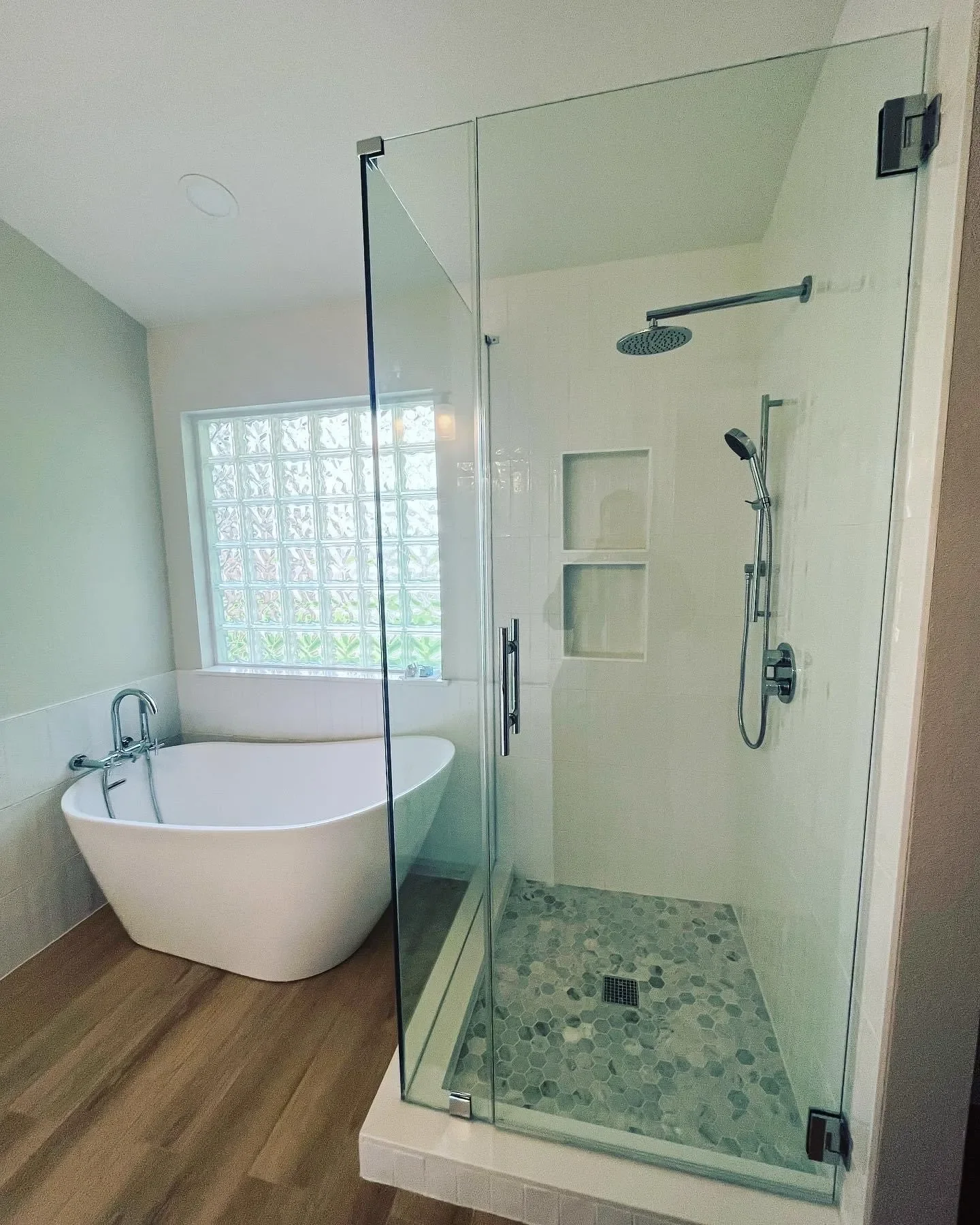 Bathroom with a white bathtub next to a window and a glass shower stall with two shower heads and tiled floor.