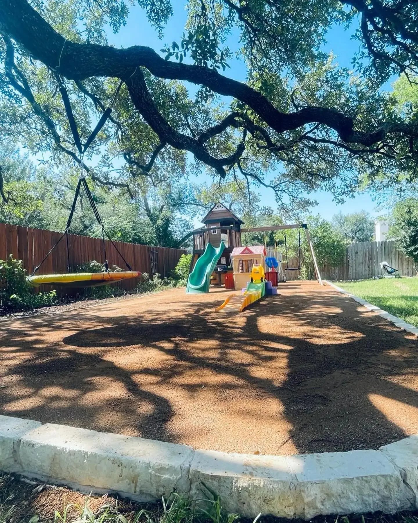 A backyard playground with swings, a slide, and a playhouse under a large tree with shade.